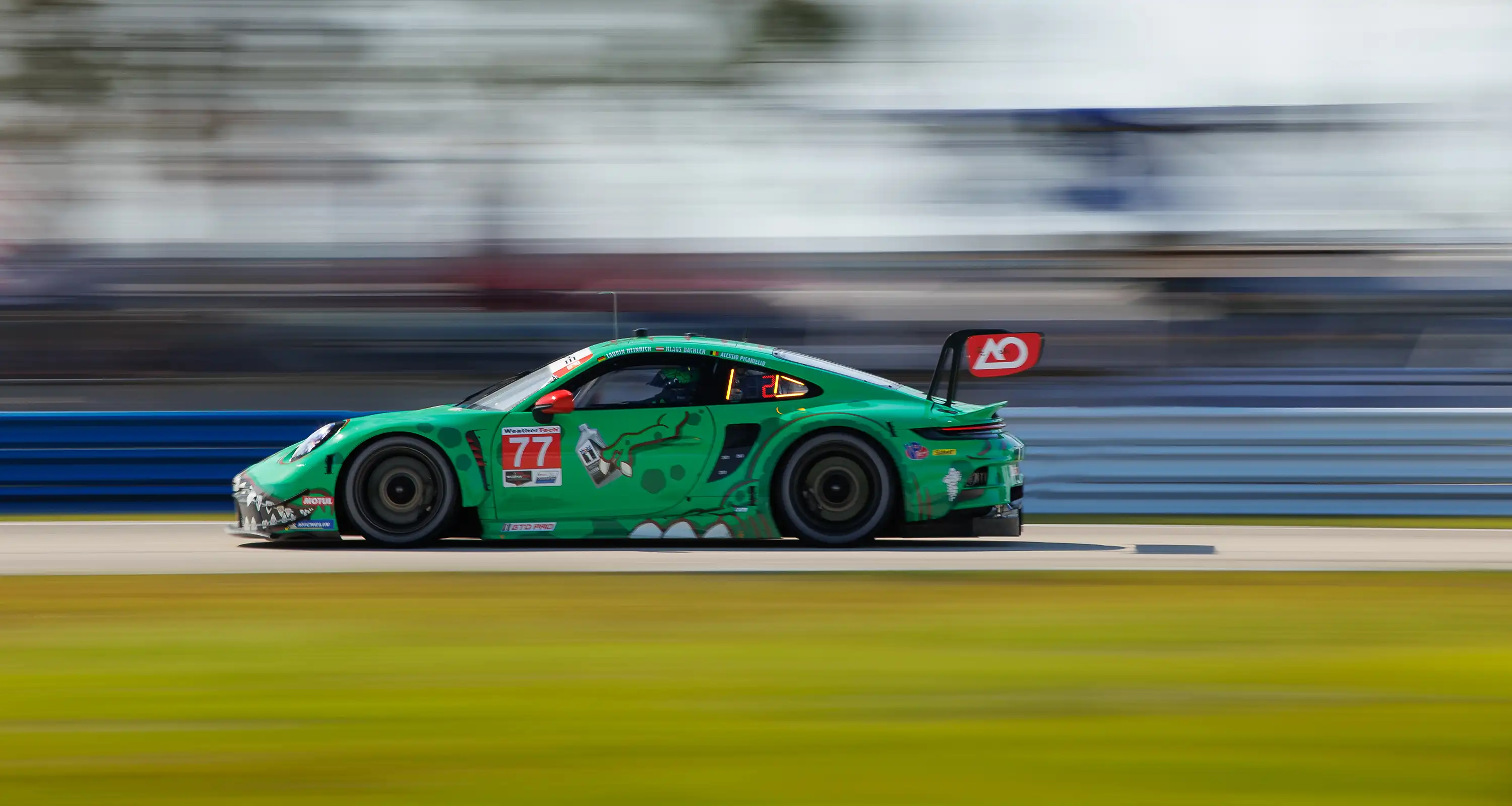A green Porsche racing car speeds around the Sebring track, with a motion-blurred background.