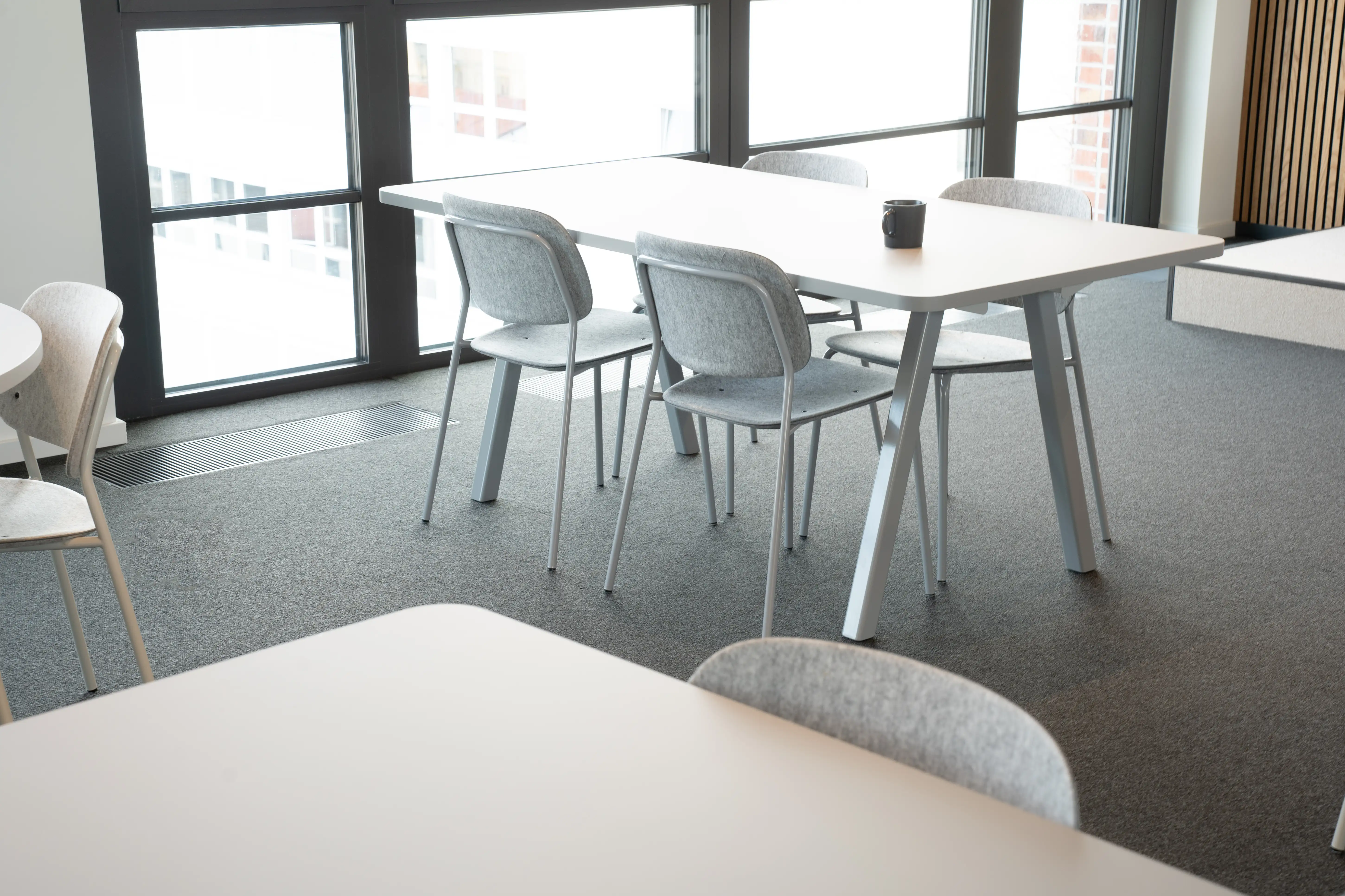 Work café tables at AI START-UP MUNICH with white tops on pale blue frames and grey felt chairs, standing on dark grey carpet by black-framed floor-to-ceiling windows.