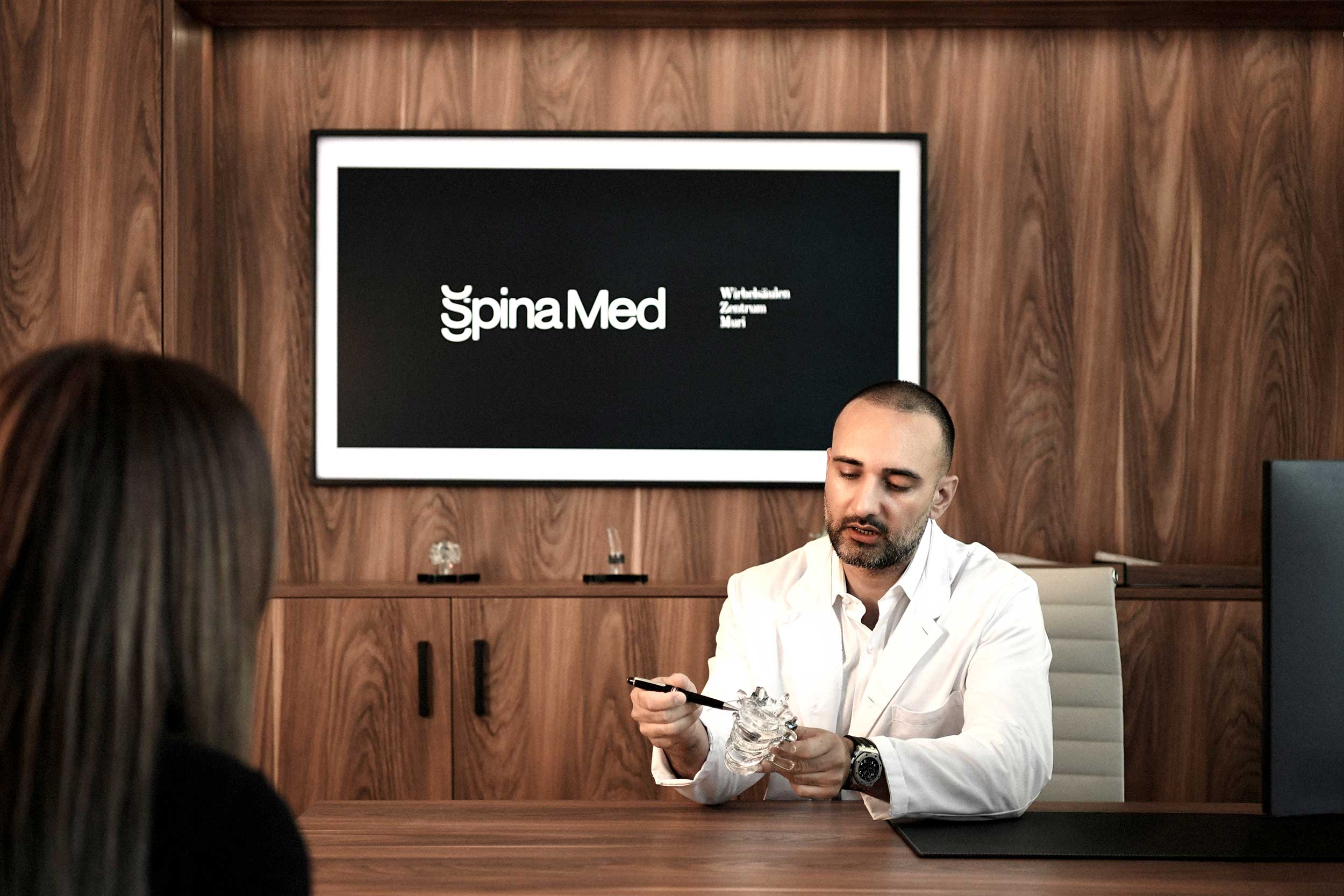 Dr. med. Sina Havakeshian erklärt einer Patientin in der Praxis SpinaMed Wirbelsäulen Zentrum Muri ein Wirbelsäulenmodell vor einem Bildschirm mit Praxislogo.