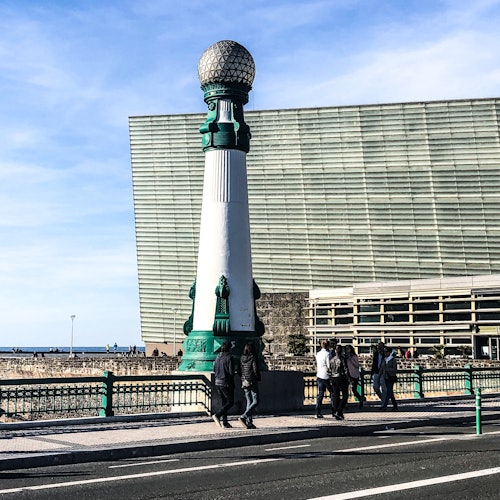 People walk by a tall, ornate green and white streetlight on a bridge, with a large modern building and the ocean in the background.