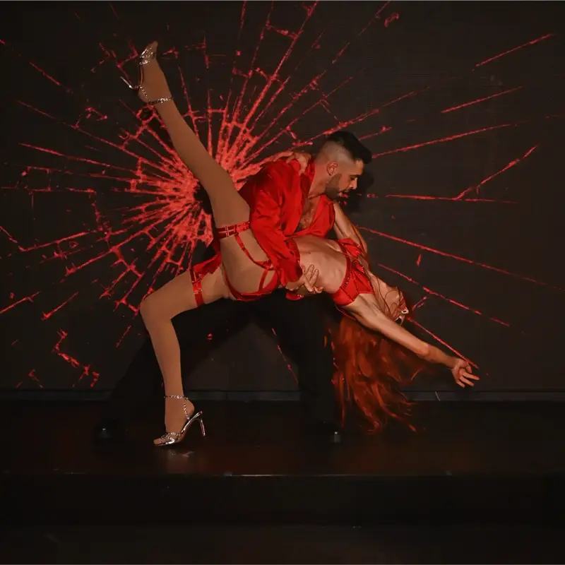 A captivating acrobatic dance performance featuring two dancers in striking red sequined costumes, with one dancer dramatically lifting and dipping another with flowing red hair against a graphic red light projection.