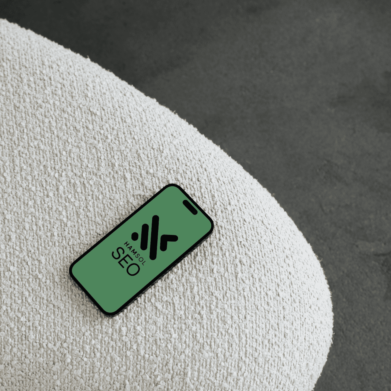 An image of an iphone laying on a white soft surface displaying the Hamilton Solutions Logo with a dark green background.