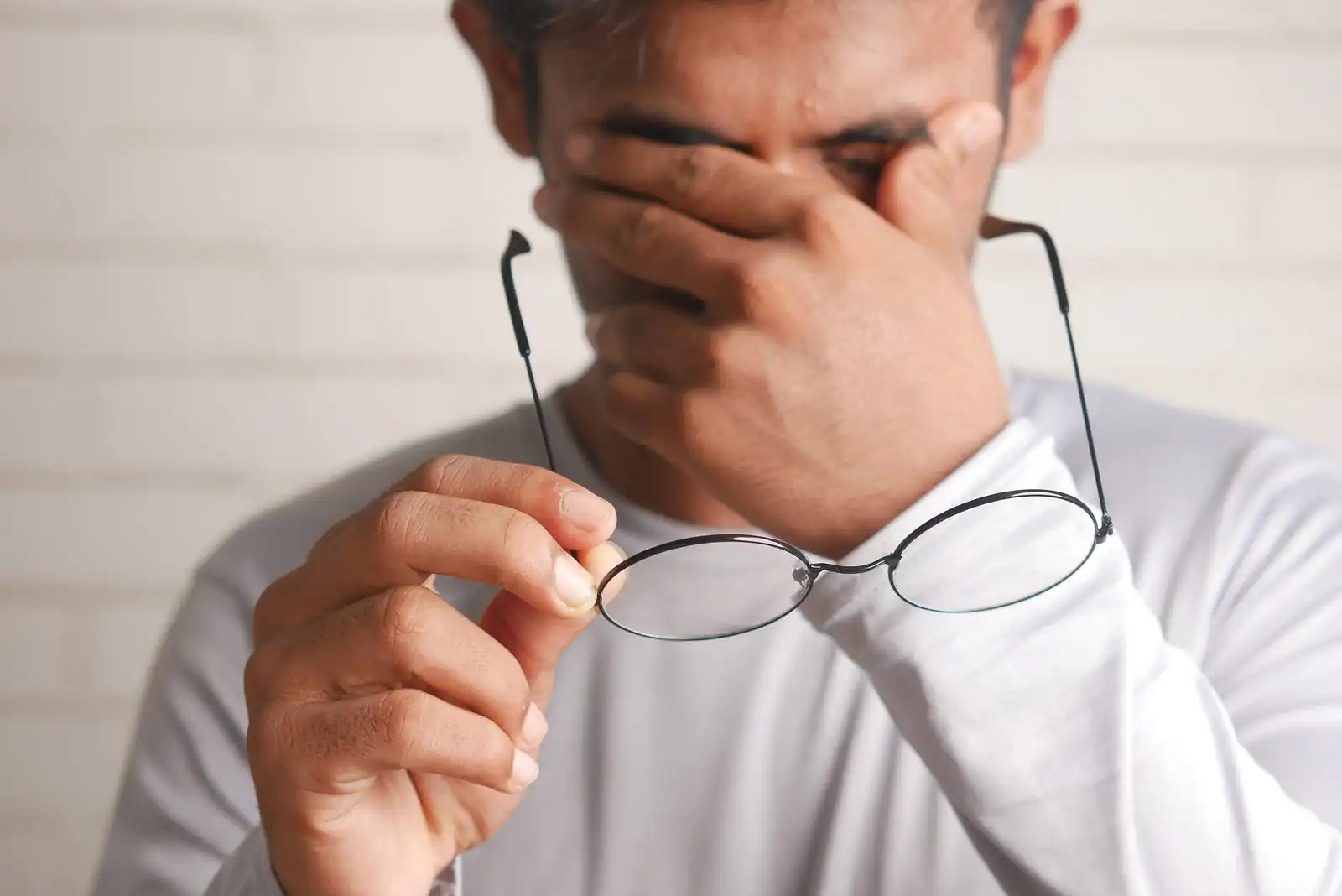 A frustrated men taking off his glasses to wipe off his face because he's so stressed. Man in white clothes in front of a white blurred wall