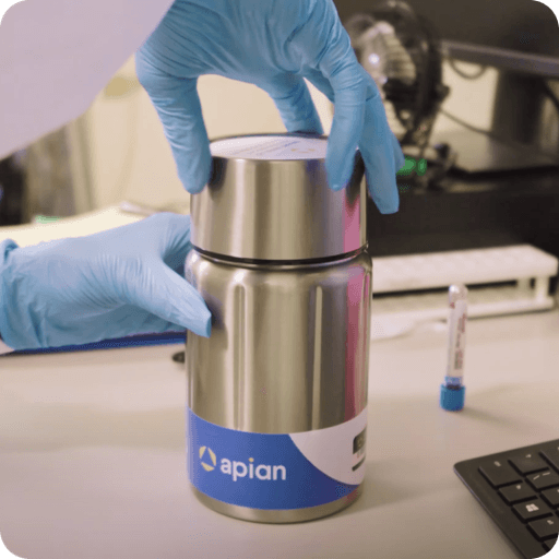 A pair of gloved hands securely closes a stainless steel container labeled "apian" on a laboratory desk, with laboratory equipment and a computer keyboard visible in the background.