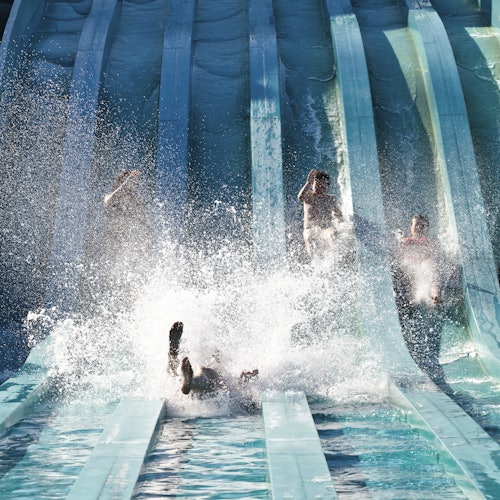 Four people slide down parallel water slides, with two splashing into the pool at the bottom.