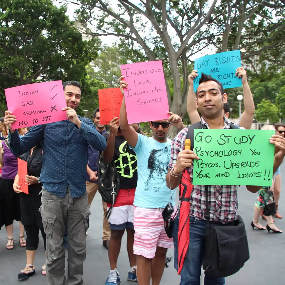 A group of people at a rally hold colourful signs supporting gay rights and protesting against outdated laws. Trees and other participants are visible in the background.