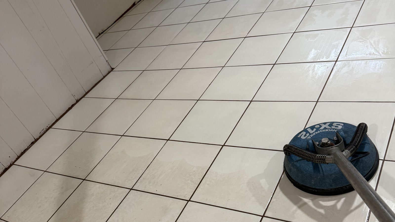 Rotary floor-cleaning tool on a white tile floor with dark grout lines.