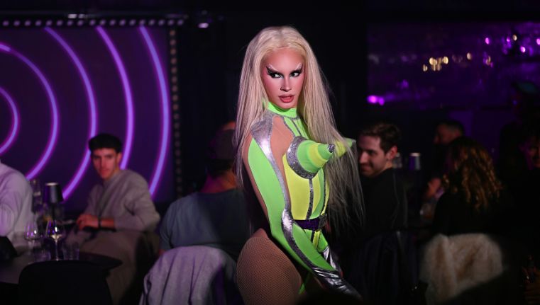 Une photo de scène dynamique d’un·e artiste drag central·e en combinaison blanche à motifs et aux cheveux blonds, tenant des bâtons néon violets lumineux, entouré·e de deux danseur·se·s de soutien