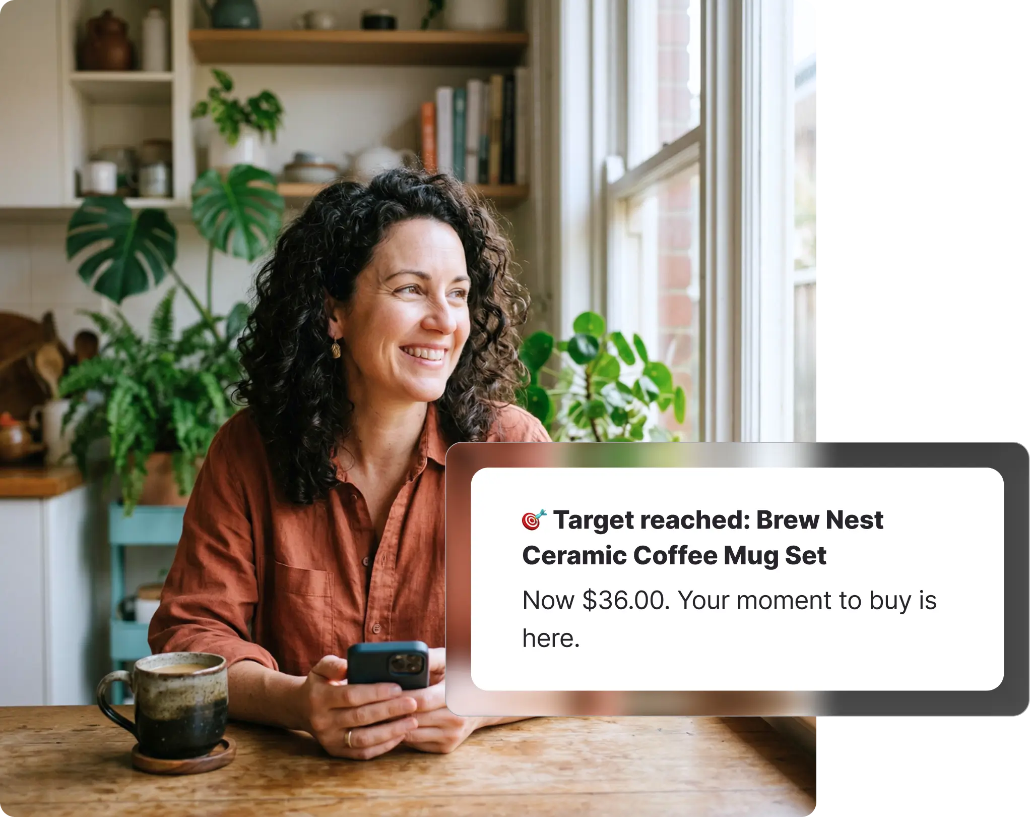 Woman with curly dark hair sitting at a kitchen table smiling at her phone, receiving a BIG W target price notification for a ceramic mug set, with indoor plants and shelves in the background.