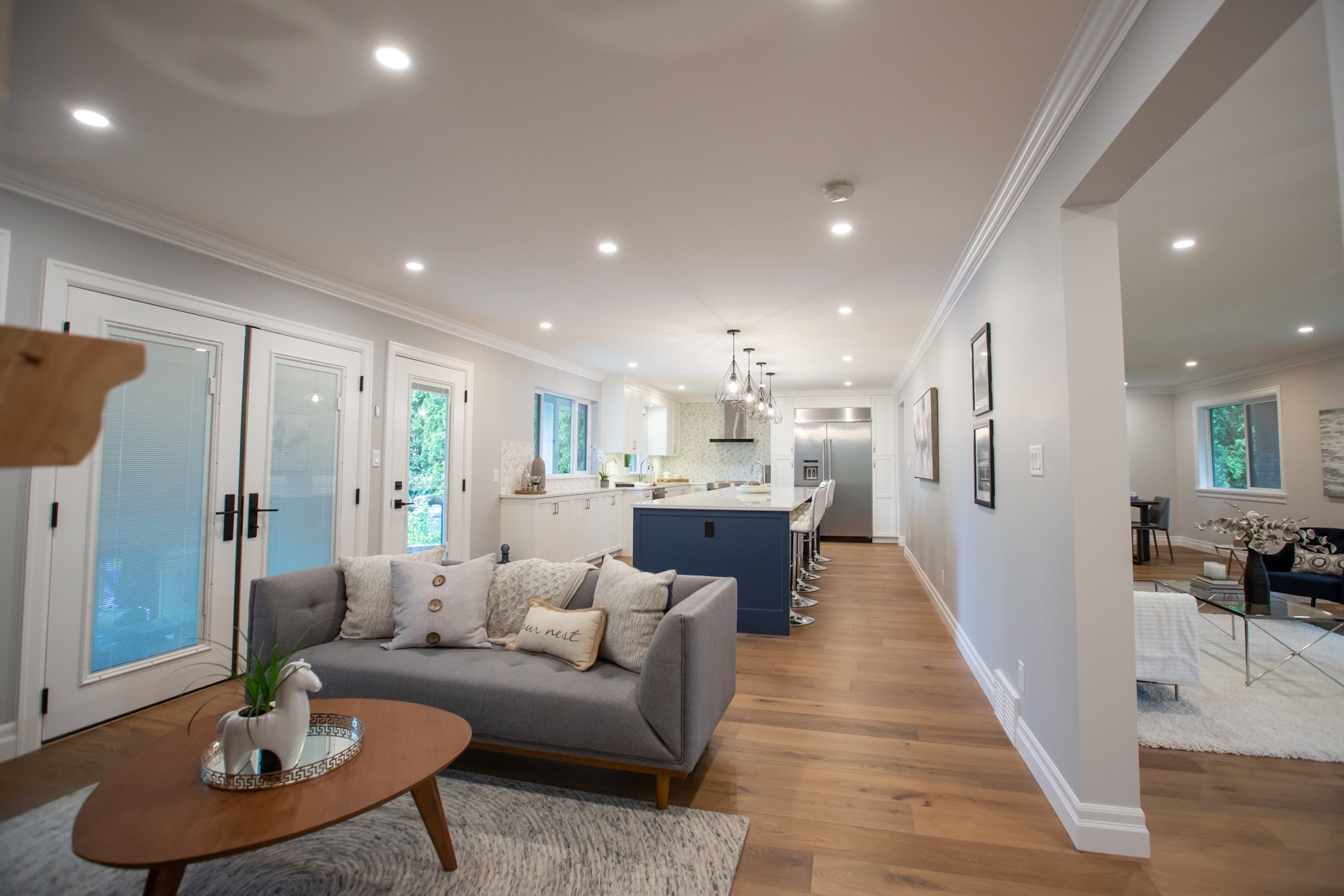 Bright, open-plan living room with a light gray sofa, round wooden coffee table, and a long kitchen island in the background under recessed ceiling lights.