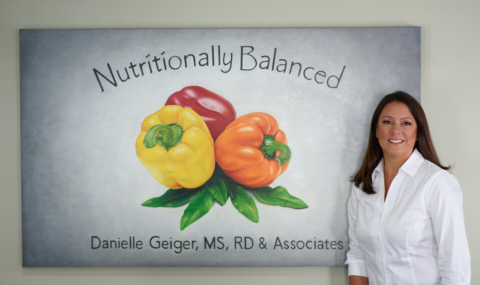 A woman in a white shirt stands next to a colorful mural featuring fruit and the text "Naturally Colored."