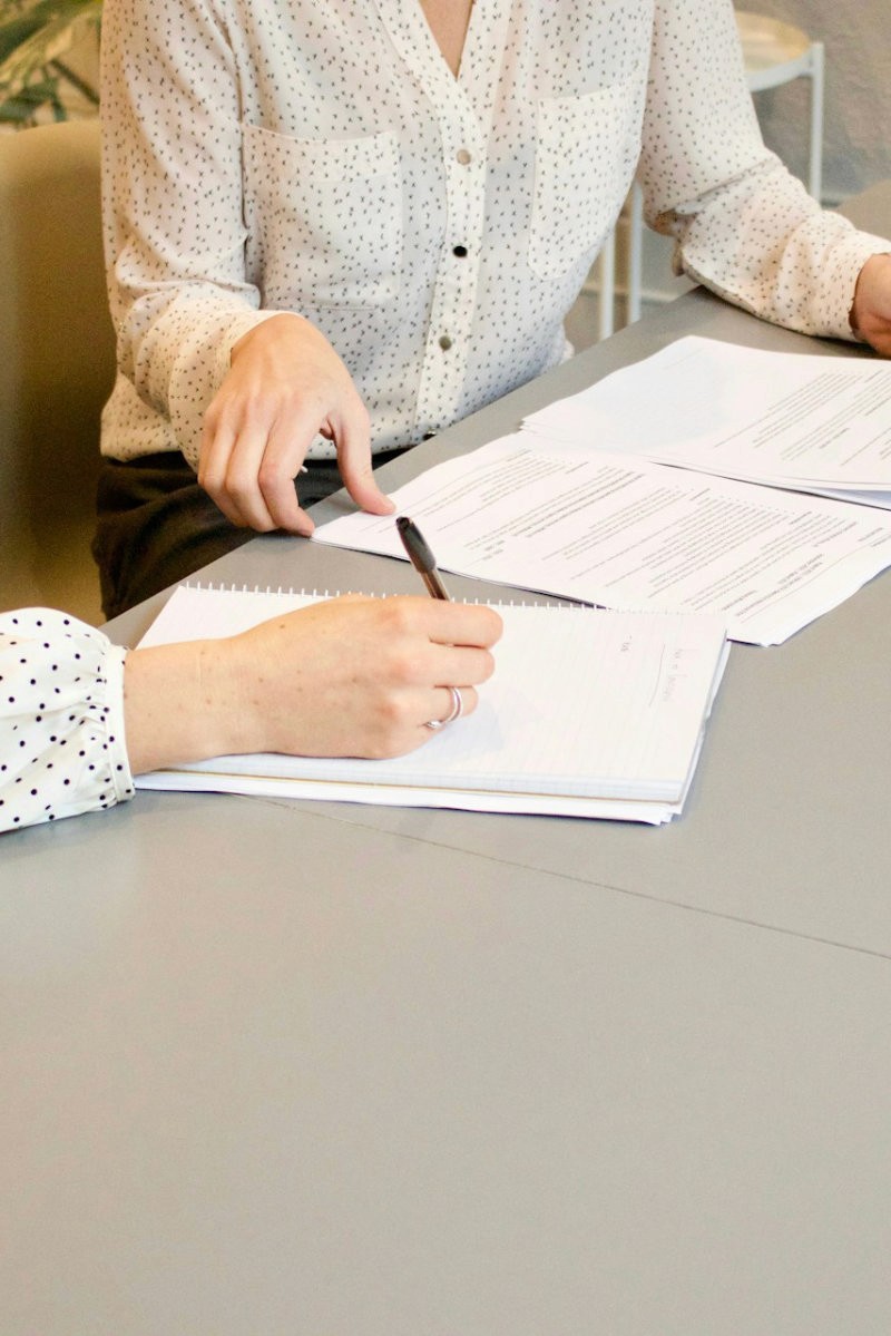 wei Frauen sitzen an einem Tisch und arbeiten gemeinsam Dokumente durch – eine schreibt, die andere zeigt auf Unterlagen