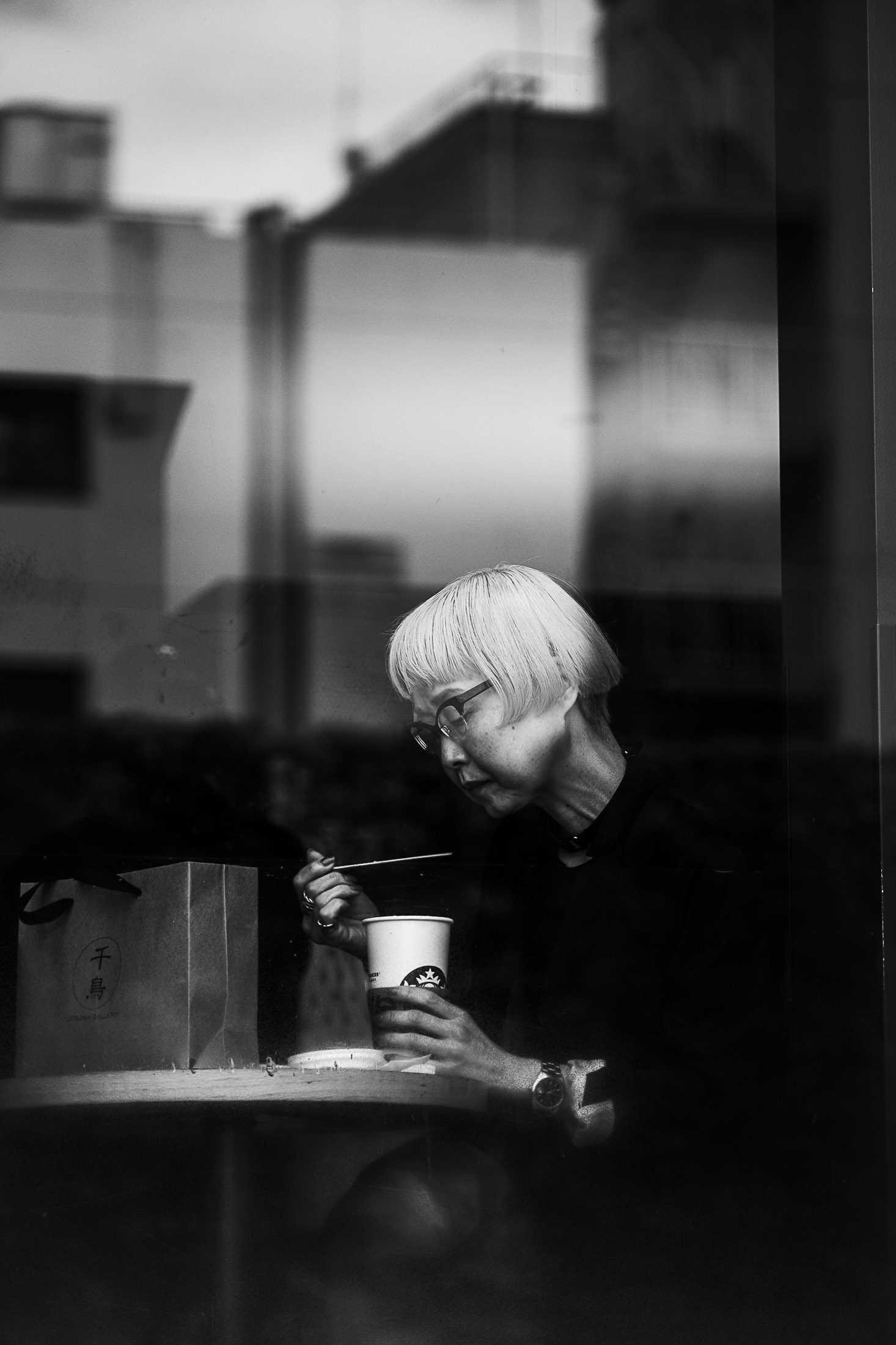 Black and white photograph of a woman eating at a small table behind a window, street photography by Richard Peterson.