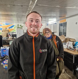 A smiling person in a black jacket stands inside a bright room filled with various items and people in the background.
