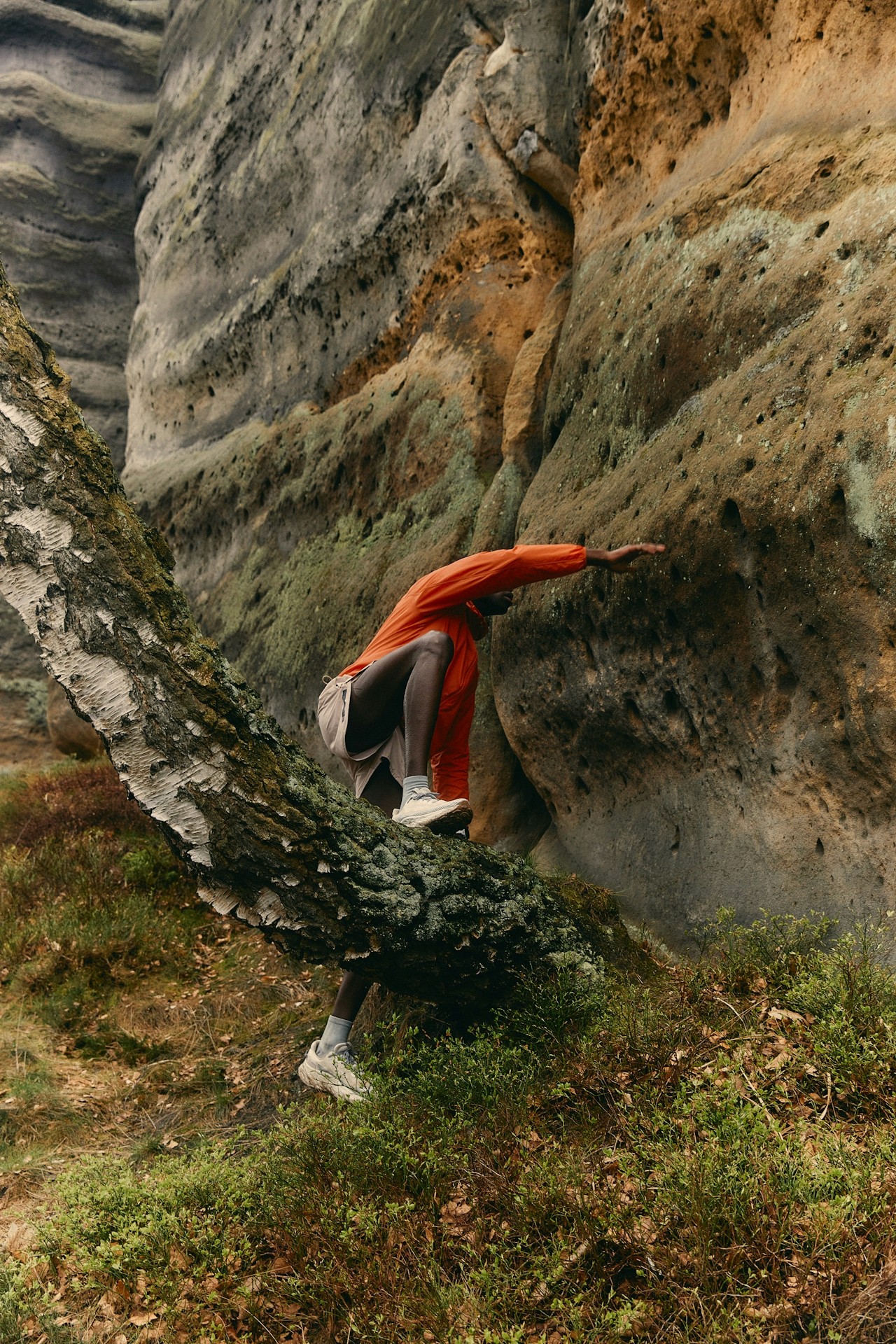 A person in an orange jacket bending near a rocky cliff in a forest.