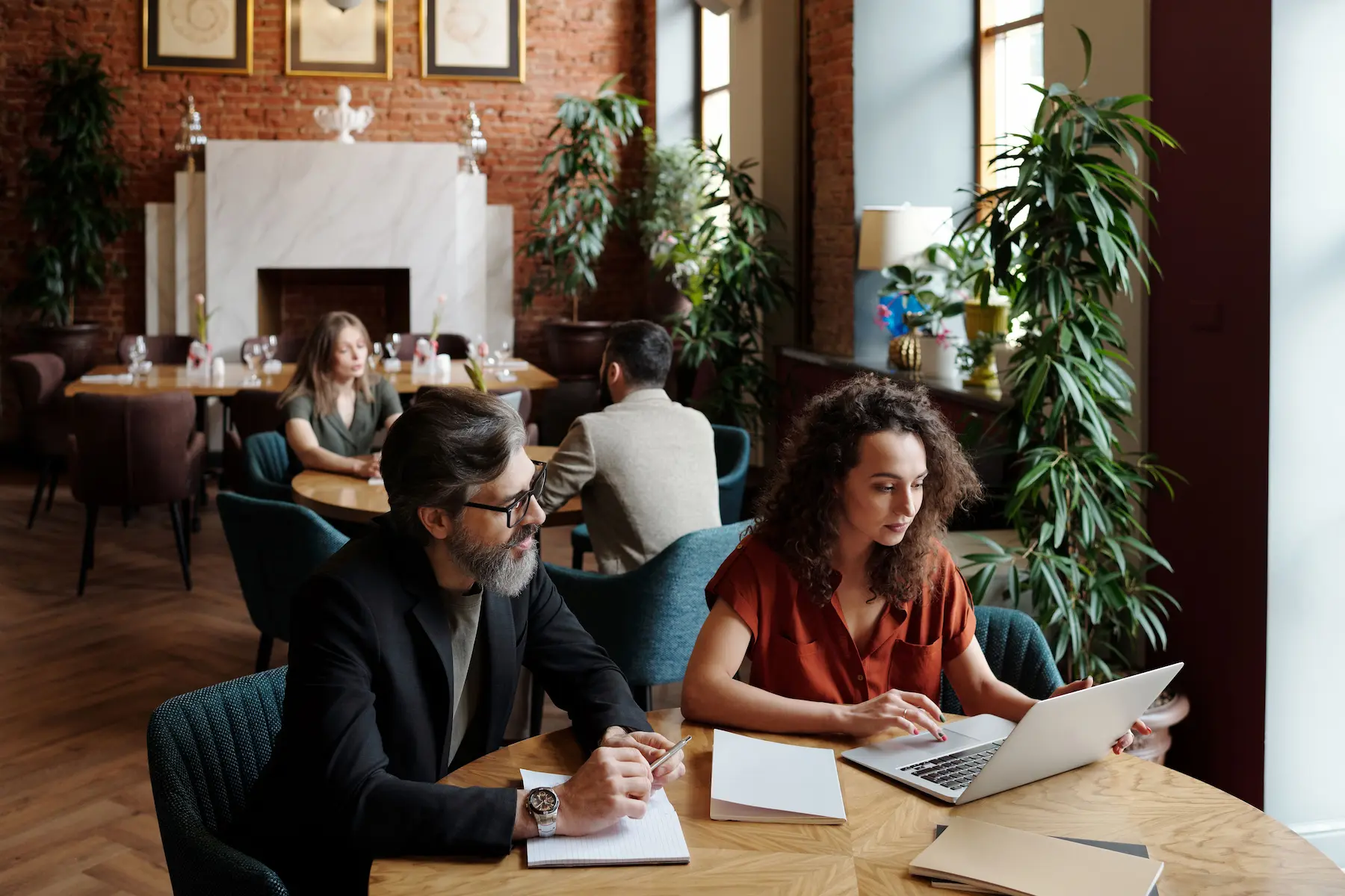 A warmly-lit cafe with brick walls and large windows features several people engaged in work, including a man and a woman sitting at a round wooden table with a laptop and notebooks, surrounded by lush green plants.