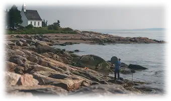 Personne installant du matériel audio sur des rochers au bord de la mer, avec une petite église blanche visible à l’arrière-plan.