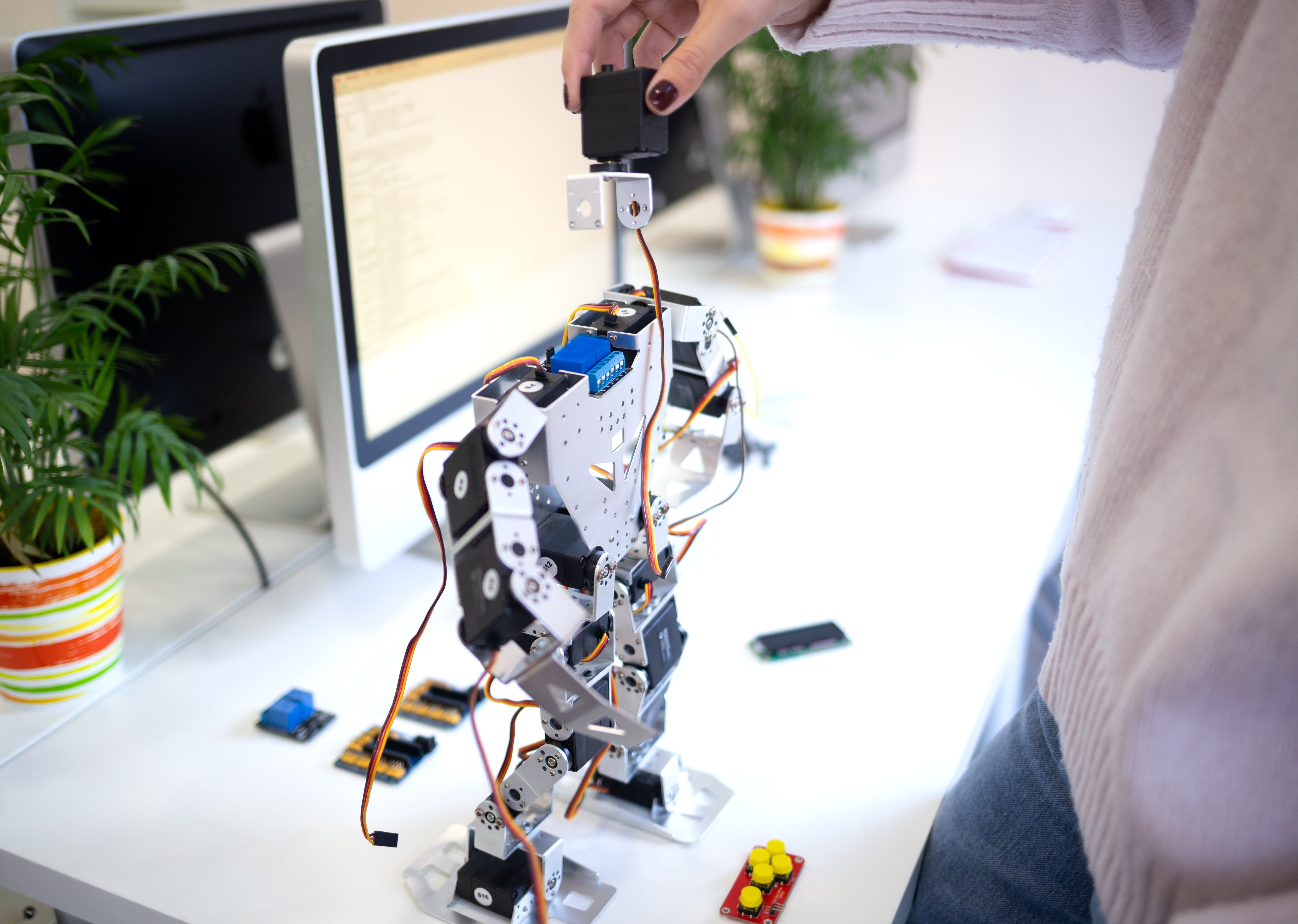 A female engineer assembles a humanoid robot on a white desk for a STEM project, with a computer showing code and electronic components in the background.
