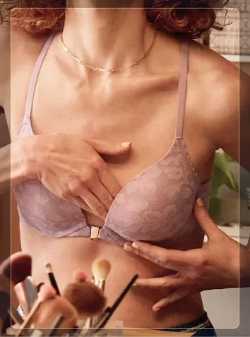 A woman in a lavender lace bra performs a breast self-exam in the mirror in a softly lit room.