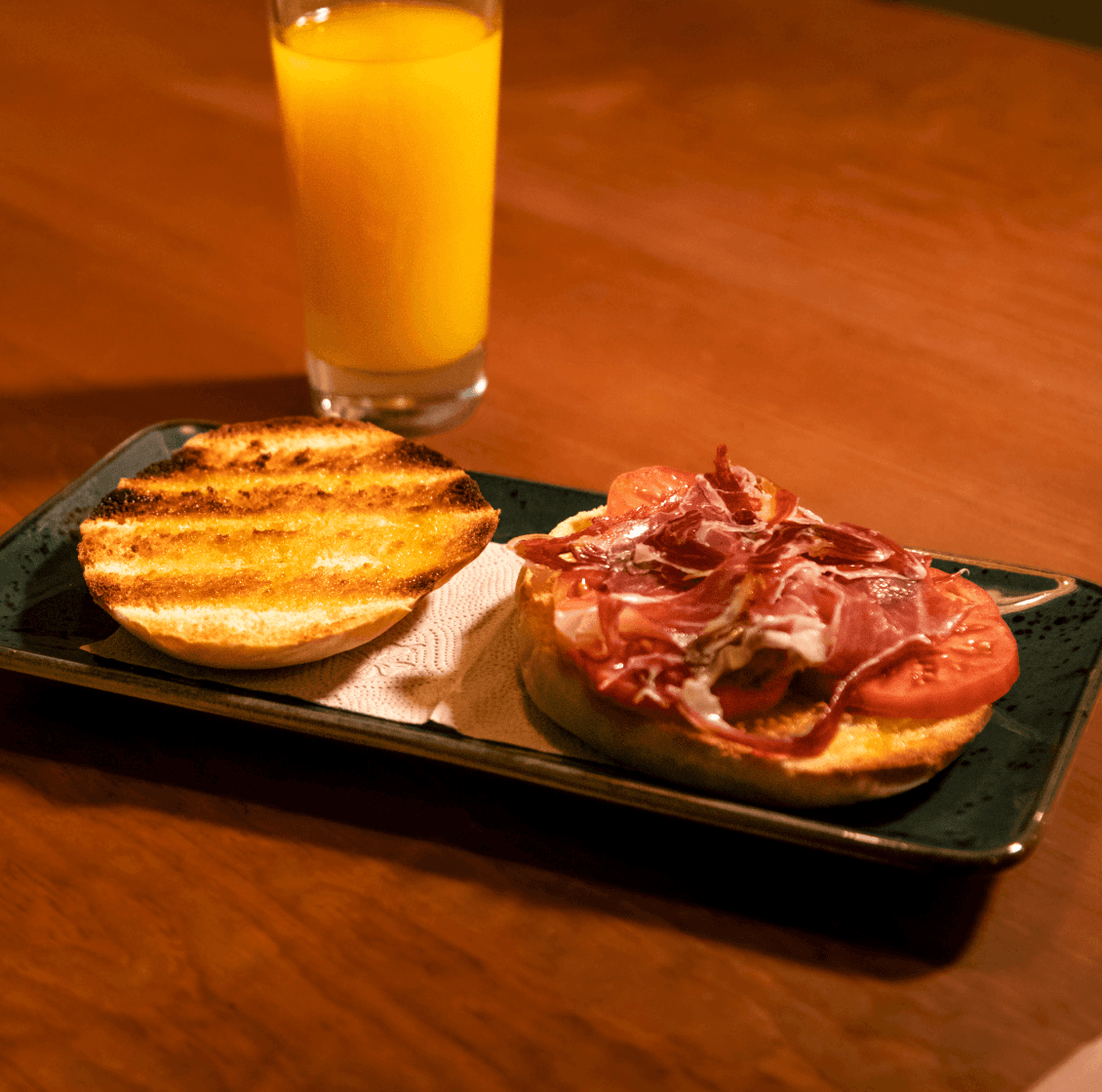 A cozy and inviting spa room with a neatly made bed, soft lighting, and warm wooden tones. The scene is partially framed by green plants, adding a natural and soothing touch.