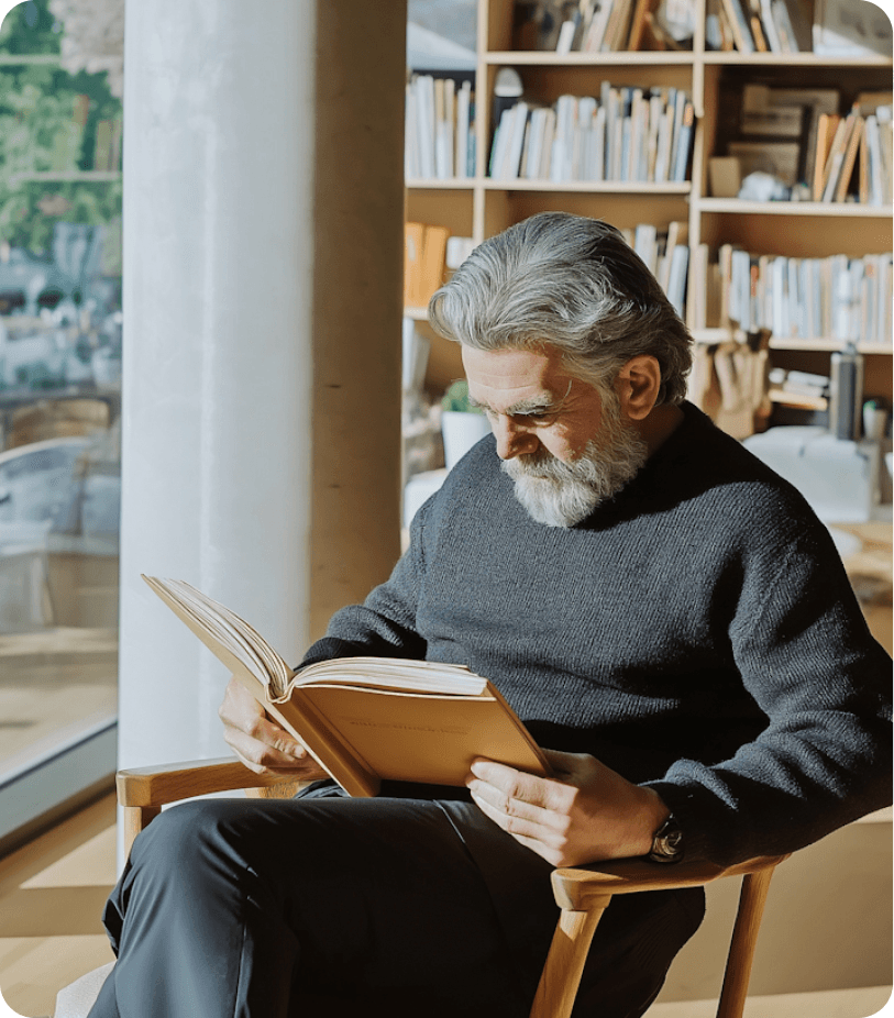 An elderly man with a gray beard, wearing a dark sweater, sits in a sunlit room with a wooden bookshelf filled with books, intently reading a book in his hands.