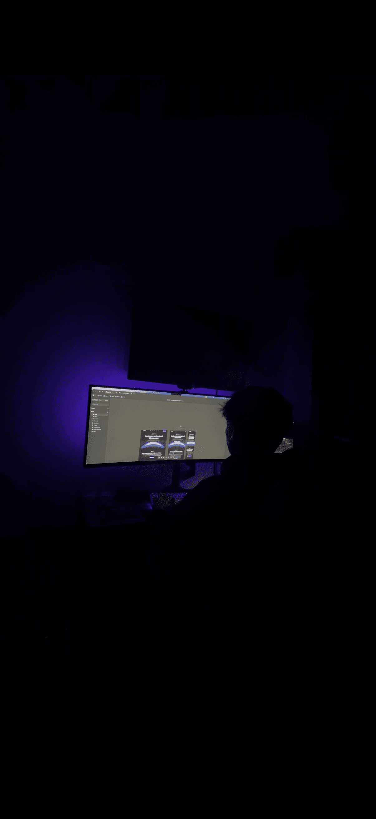 A person is sitting in a dark room at a desk, working on a dual-monitor setup with a purple glow illuminating the space, creating a focused and tech-savvy ambiance.
