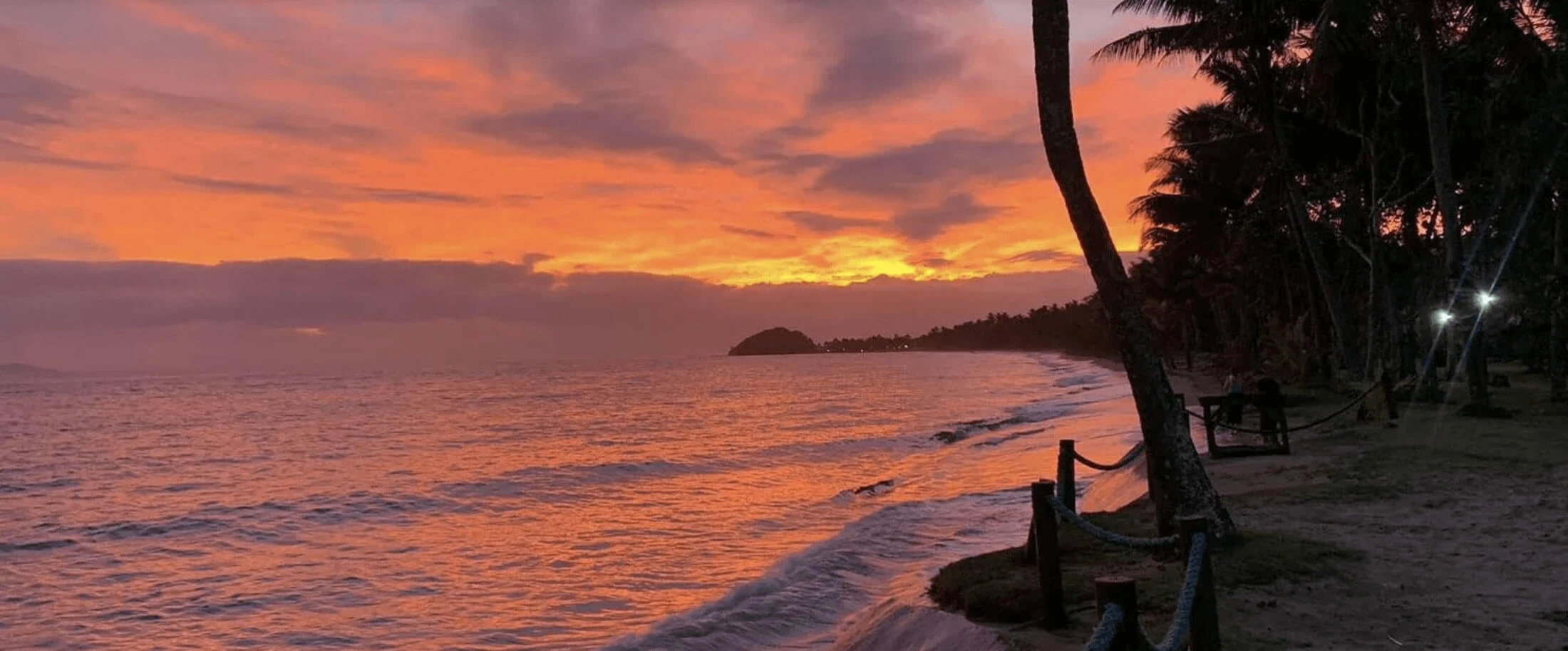 Golden hour sunset image of sandy beaches in Fiji, palm tree pictured right, as the waves crash gentley on the shoreline, the sun is setting