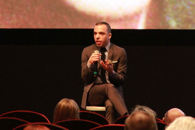A man in a dark suit and tie sits on a stool, holding a microphone and gesturing with his hands. He faces the audience, seated in a row of red chairs. A large screen behind him displays a blurred image. The setting appears to be a conference or presentation hall.