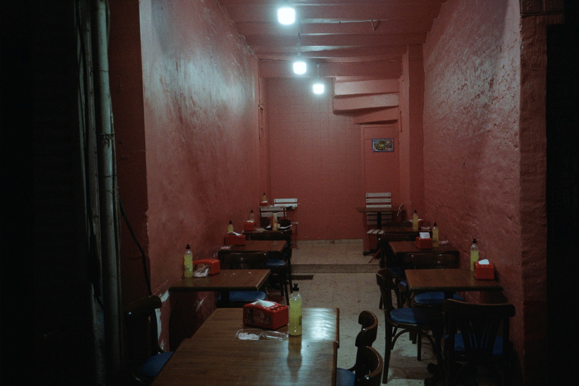 A dimly lit, narrow restaurant interior with pink walls features wooden tables and chairs, each set with bottled condiments and red napkin holders, creating a cozy, intimate dining atmosphere.