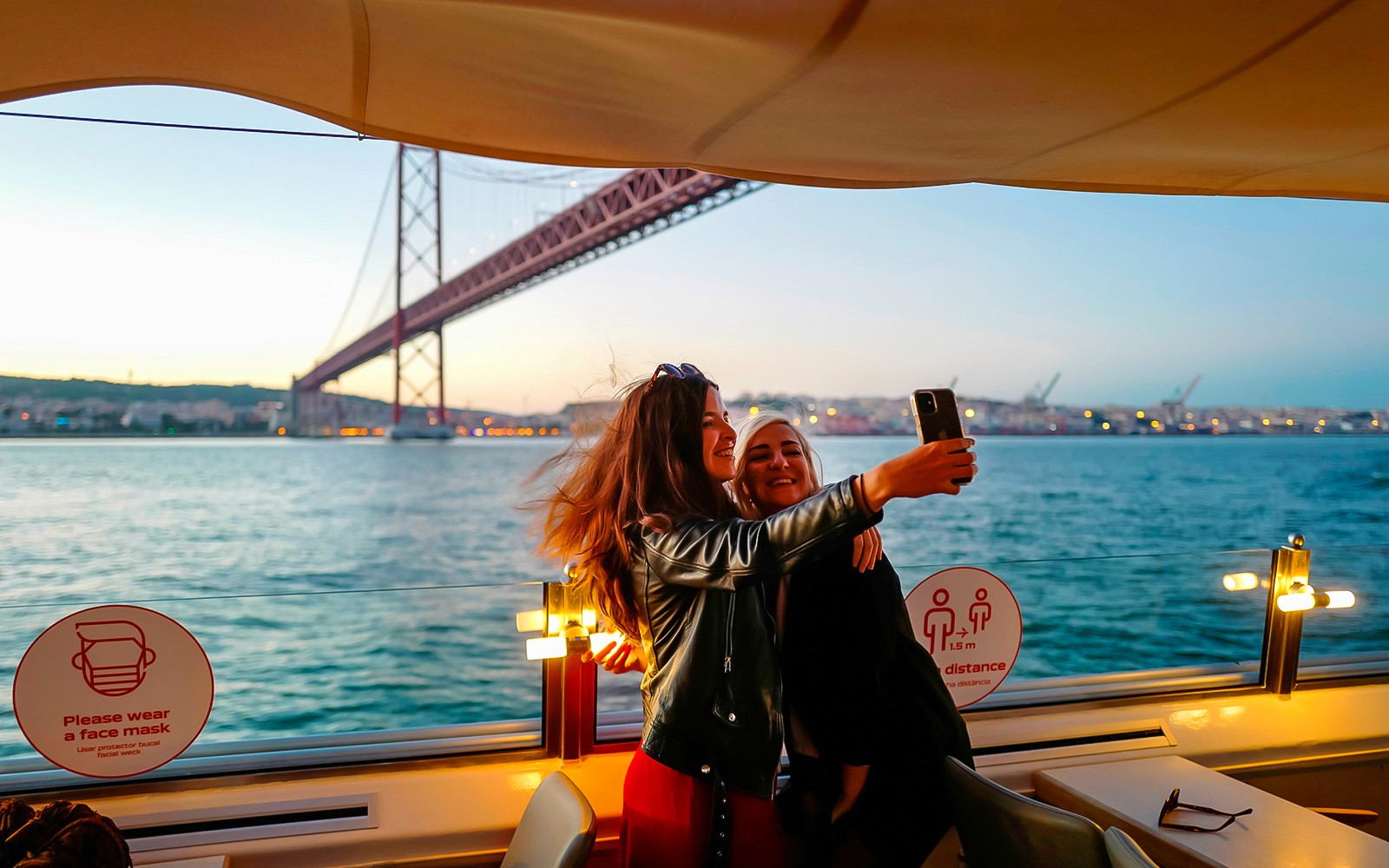 Dwie osoby robiące sobie selfie podczas rejsu o zachodzie słońca po rzece Tag w Lizbonie, z mostem w tle.