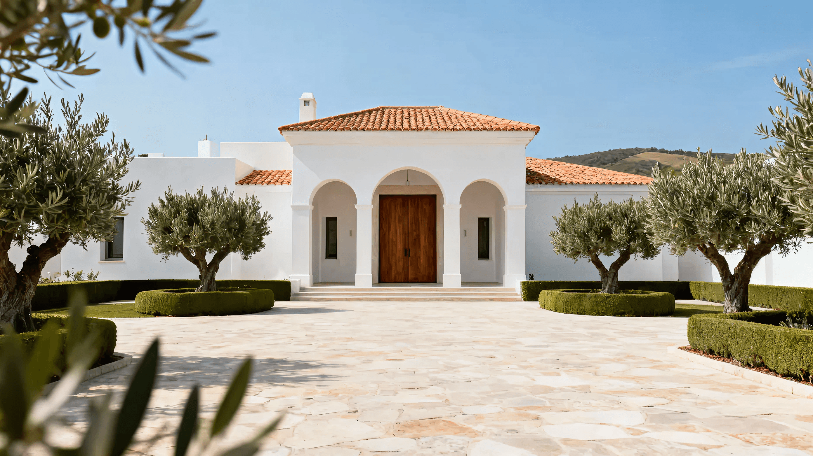 Mediterranean-style villa with white walls, terracotta roof, arched windows, and a stone pathway lined with potted olive trees.