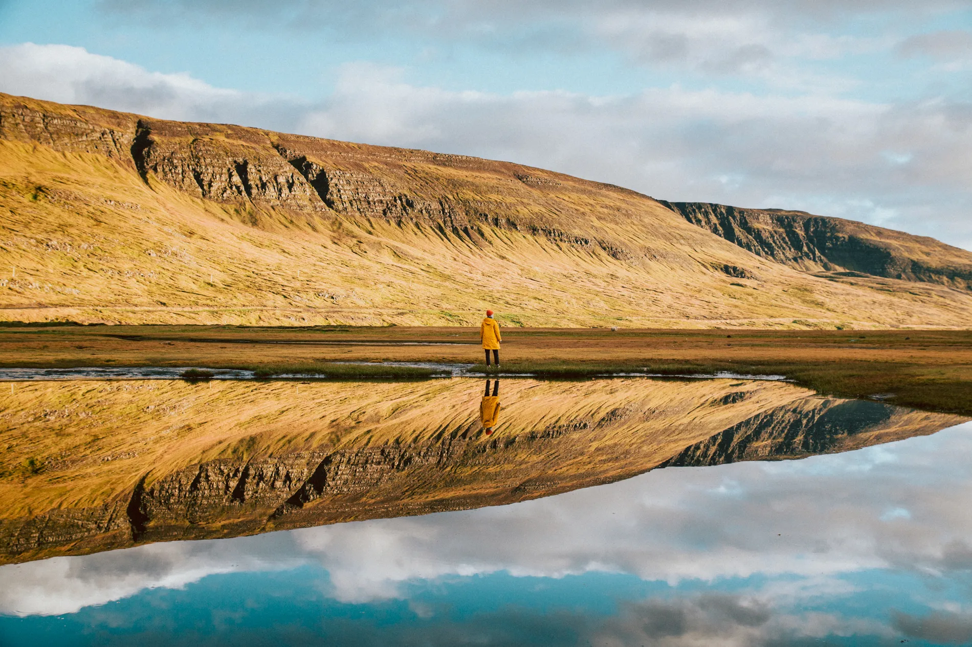 A person in yellow standing at the edge of a mirror-still lake below golden cliffs
