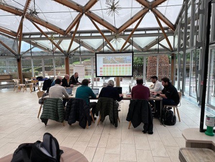 A group of people seated around a table in a glass-enclosed room, watching a presentation on a screen.
