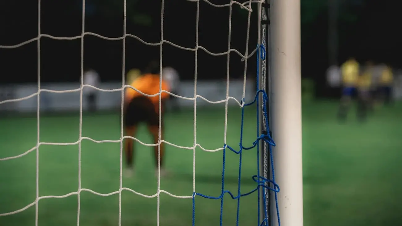 Close-up view of a football net with a goalkeeper and players in the background during an amateur league match, highlighting the grassroots aspect of the sport which is the focus of the Baller League, aiming to bridge traditional football with modern entertainment.