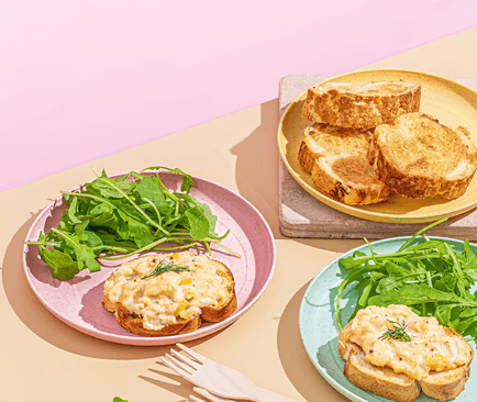 Creamy egg products on toasted bread with fresh green arugula, served on pastel pink and mint plates under bright sunlight.