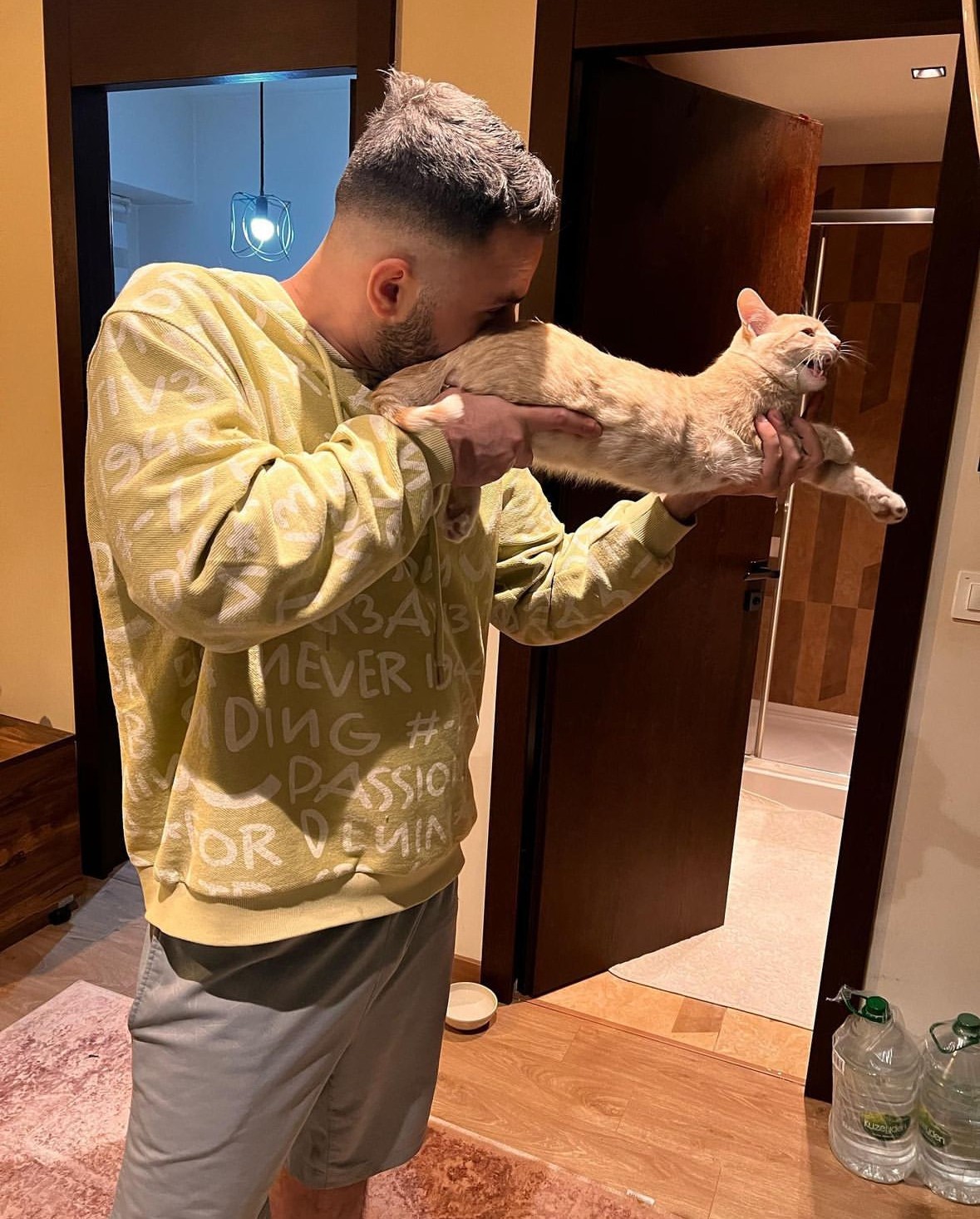 Man wearing a mustard-yellow sweatshirt gently kisses an orange tabby cat in a doorway, capturing an affectionate moment.