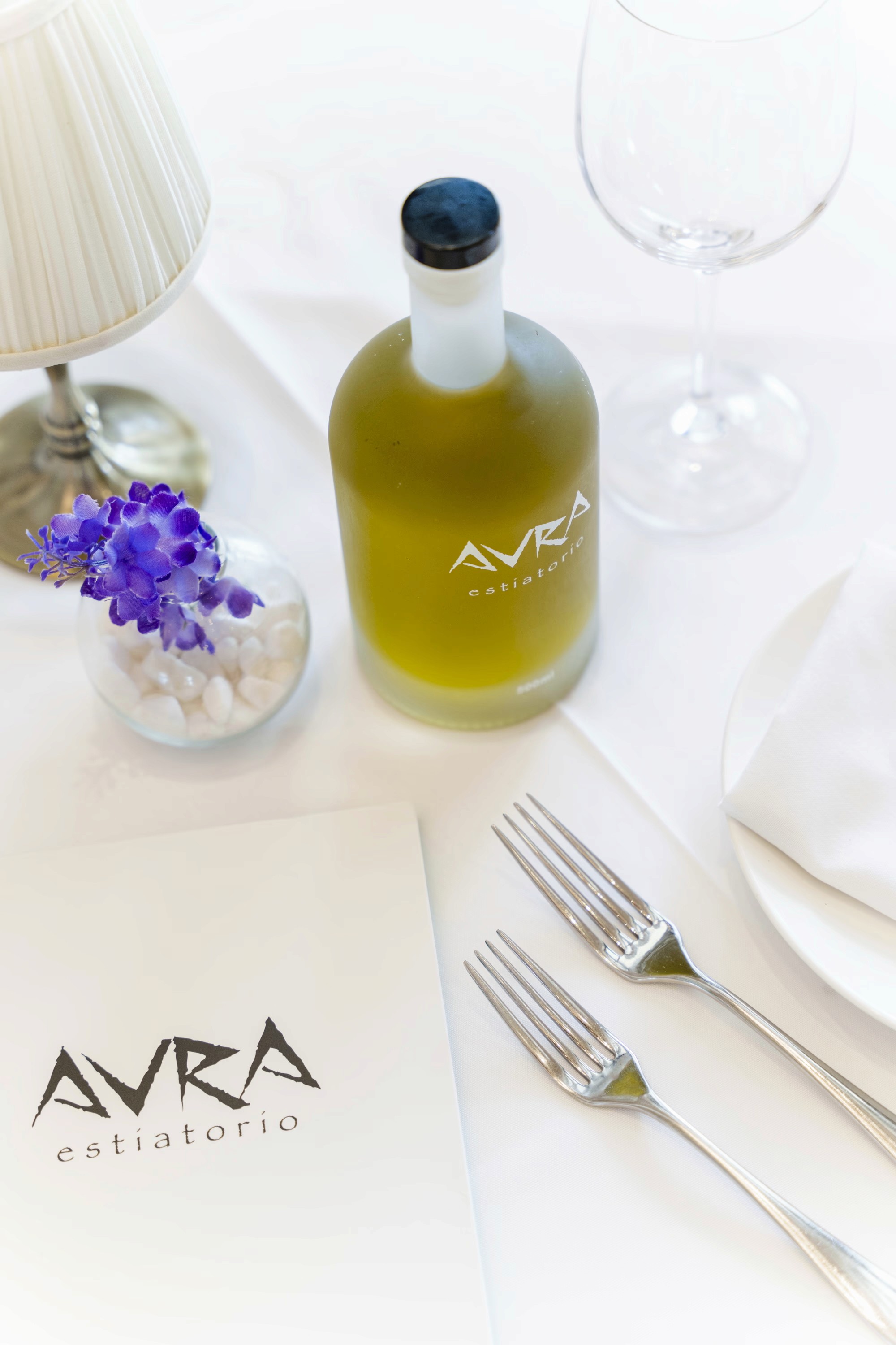 A glass bottle of olive oil with a purple label next to utensils and a folded napkin on a white surface.