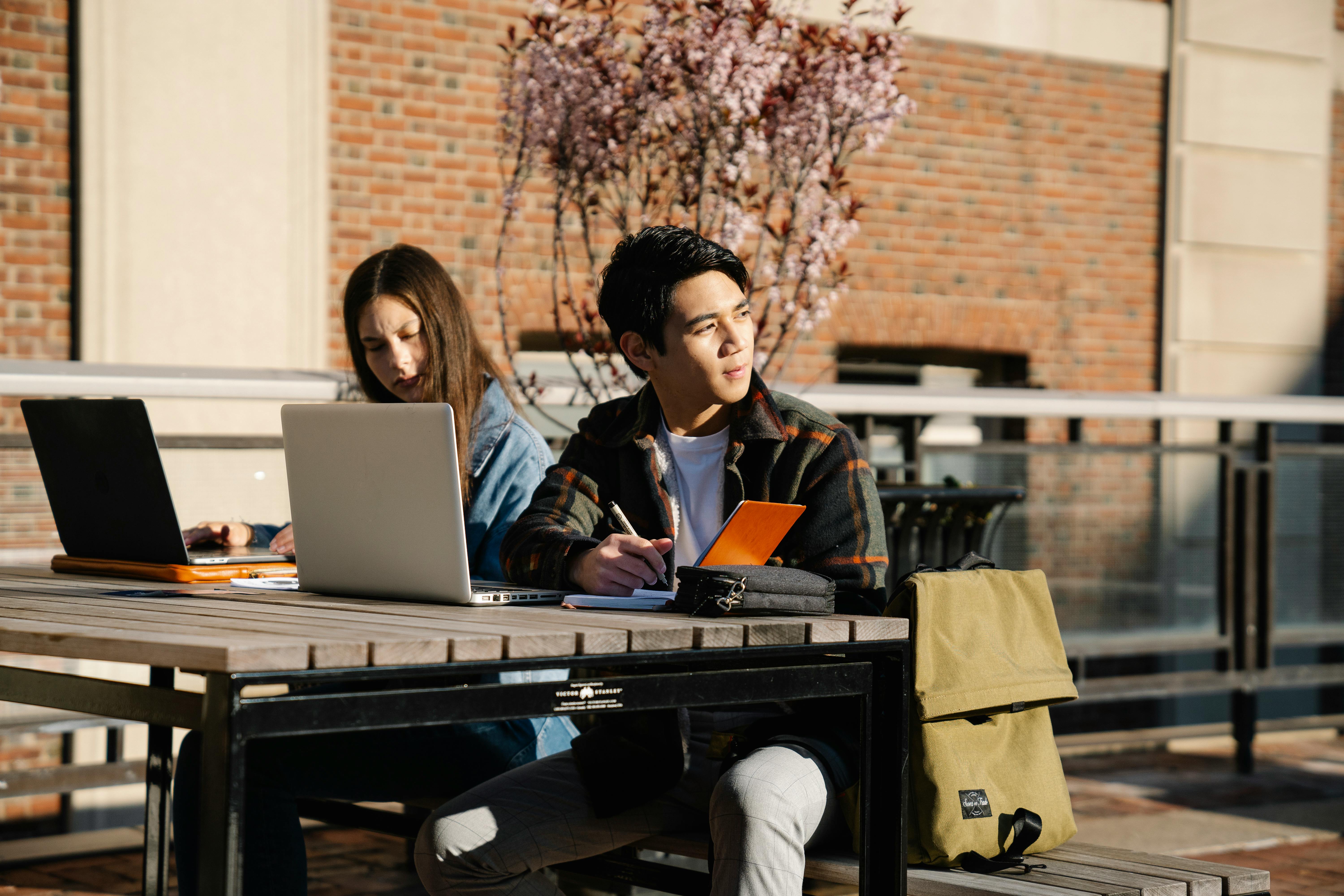 Two university students are sitting outdoors at a wooden table on an urban Canadian campus. A woman with long brown hair, wearing a denim jacket, is focusing on a dark grey laptop. Beside her, a man of East Asian descent, wearing a plaid shirt over a white tee, holds a notebook and pen, looking off to the side. Both of their laptops are visible, one being a silver model. A green canvas backpack sits on the ground next to them. This image captures the authentic Canadian student experience and the necessity of a reliable, durable laptop for studying outdoors, even as winter approaches.