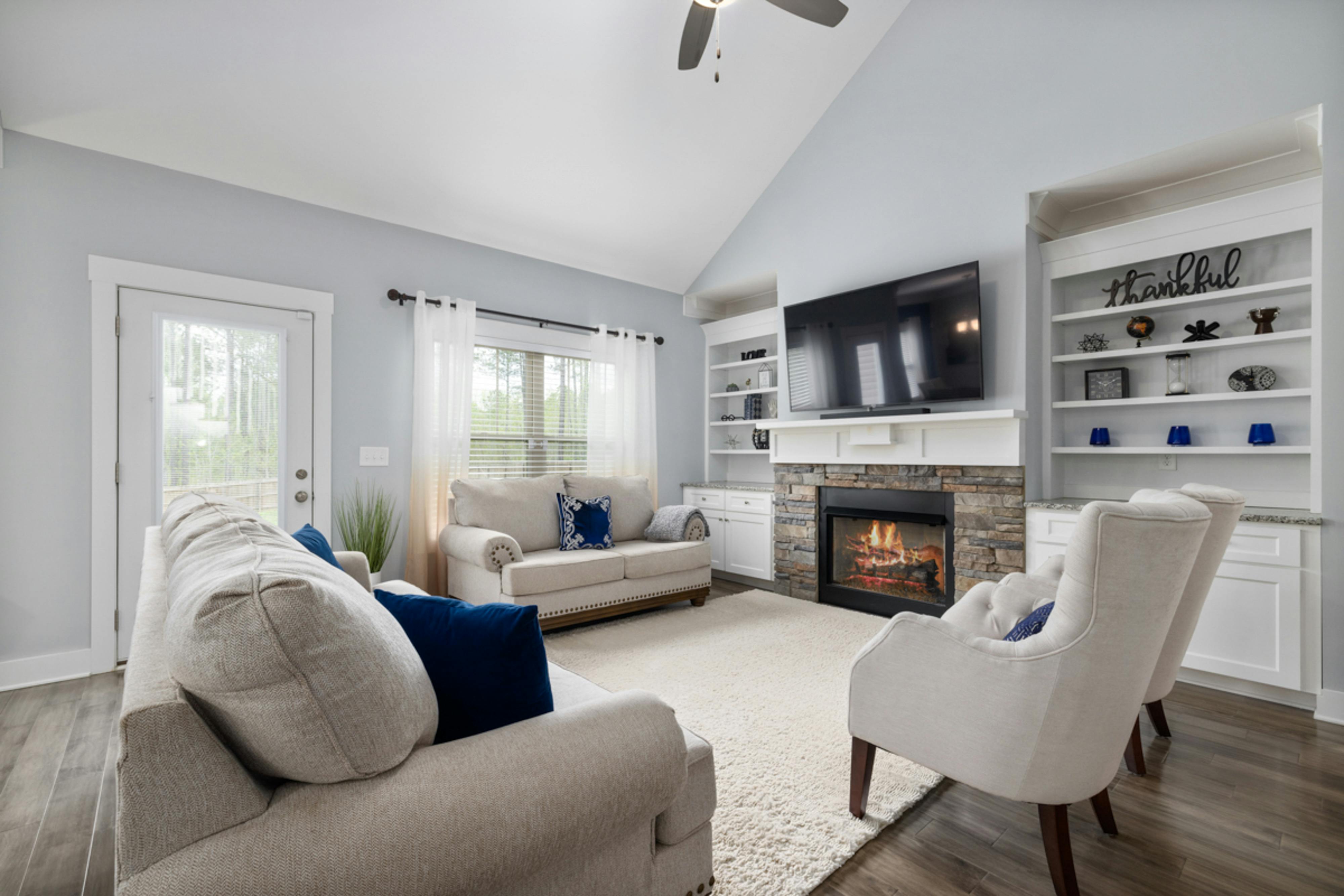 Bright and spacious living room with modern furnishings, featuring a stone fireplace, wall-mounted TV, and built-in shelves