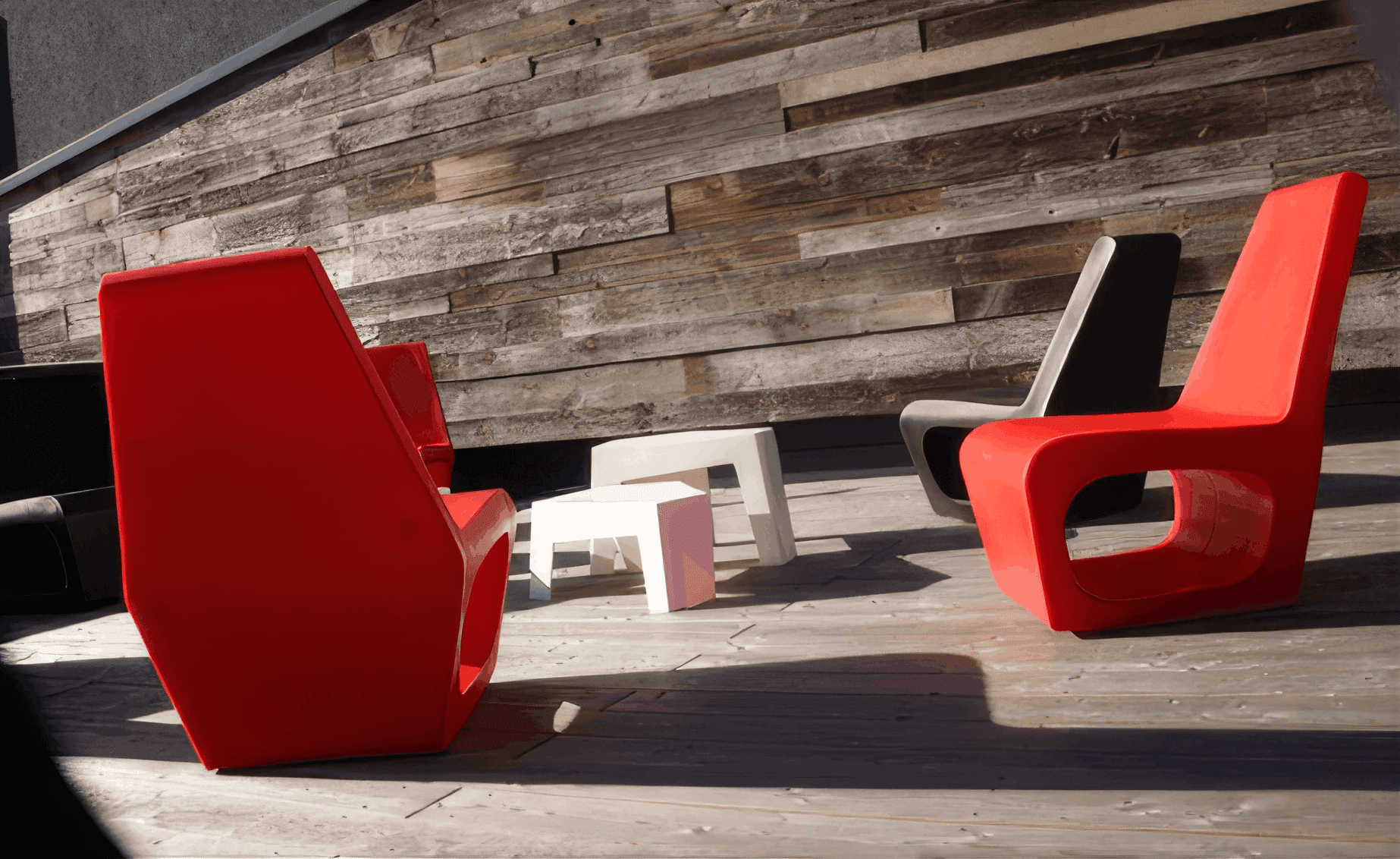 MW Interior  - Zwei rote Designsessel aus Kunststoff mit kleinem weißem Beistelltisch auf Holzdeck vor rustikaler Altholzwand" Beschreibung: Markante Outdoor-Lounge mit wetterbeständigen Designsesseln in Signalrot auf naturbelassenem Holzdeck – der Kontrast zur verwitterten Altholzwand macht die Situation zum gestalterischen Statement.