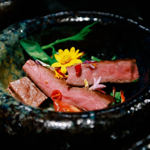 Felii de carne de vită gătită, garnisite cu flori comestibile și verdețuri, servite într-un bol de ceramică închisă la culoare.