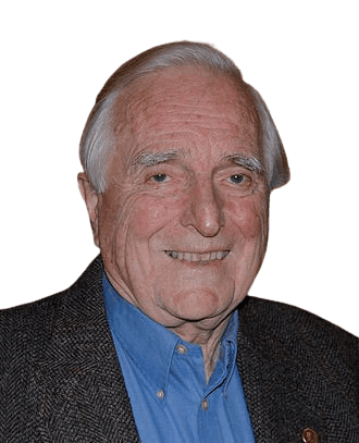 An older man with gray hair and a friendly smile is pictured indoors, wearing a blue collared shirt and a dark textured blazer. His warm expression and relaxed appearance give the portrait an approachable and amiable feel, suitable for depicting an experienced professional or distinguished figure in a social setting.