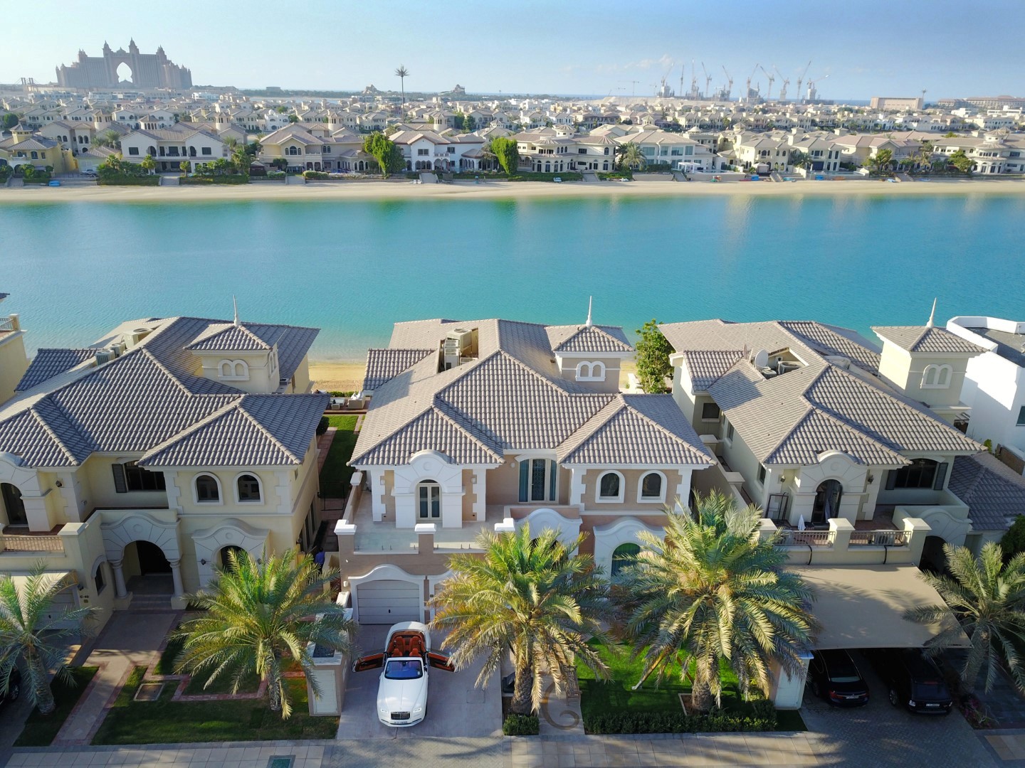 Aerial view of luxury waterfront villas with tiled roofs and palm trees, situated along a tranquil blue waterway, under a clear sky with a city skyline in the background.