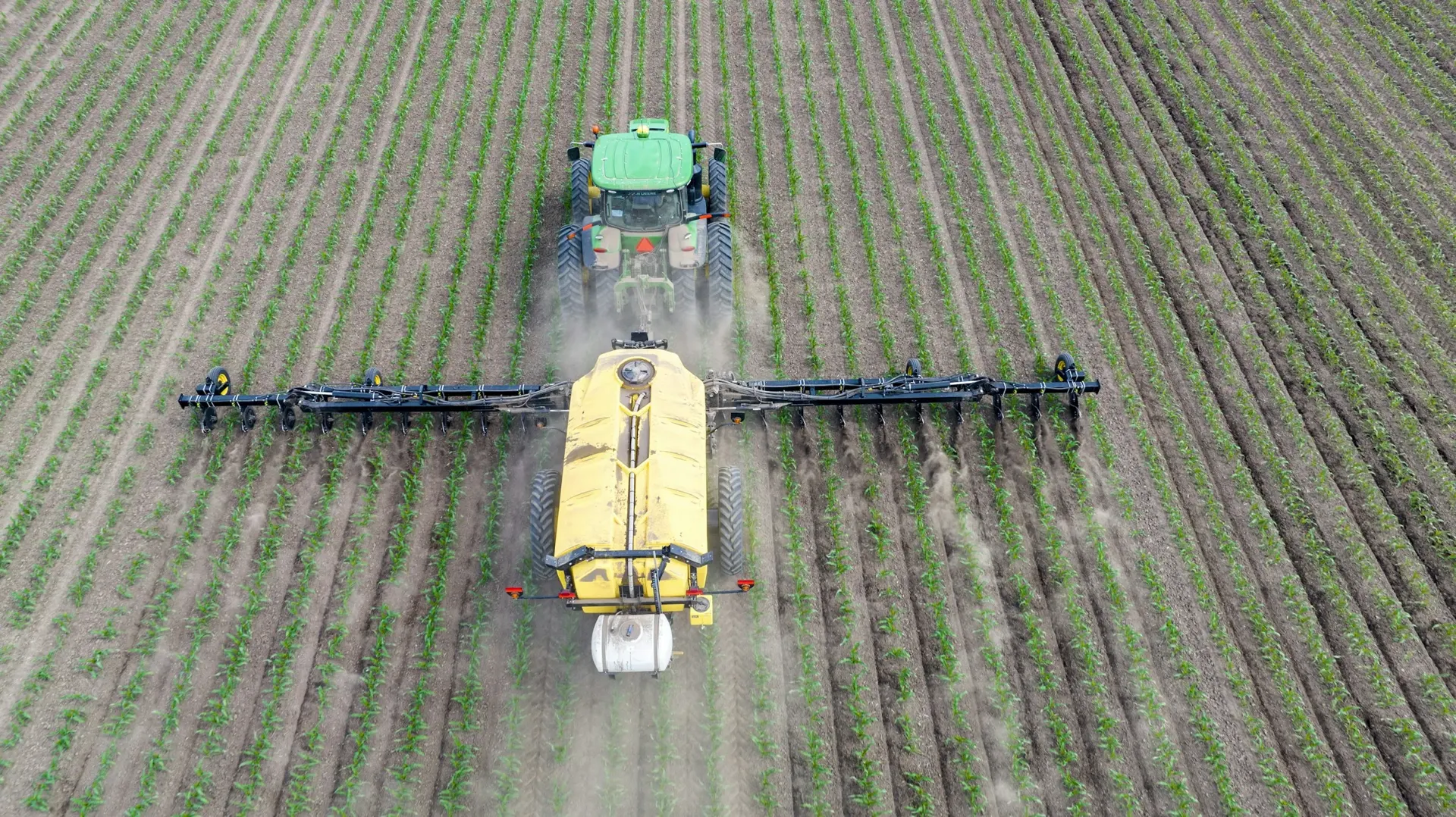 Aerial view of a tractor applying fertilizer across rows of young crops in a green field
