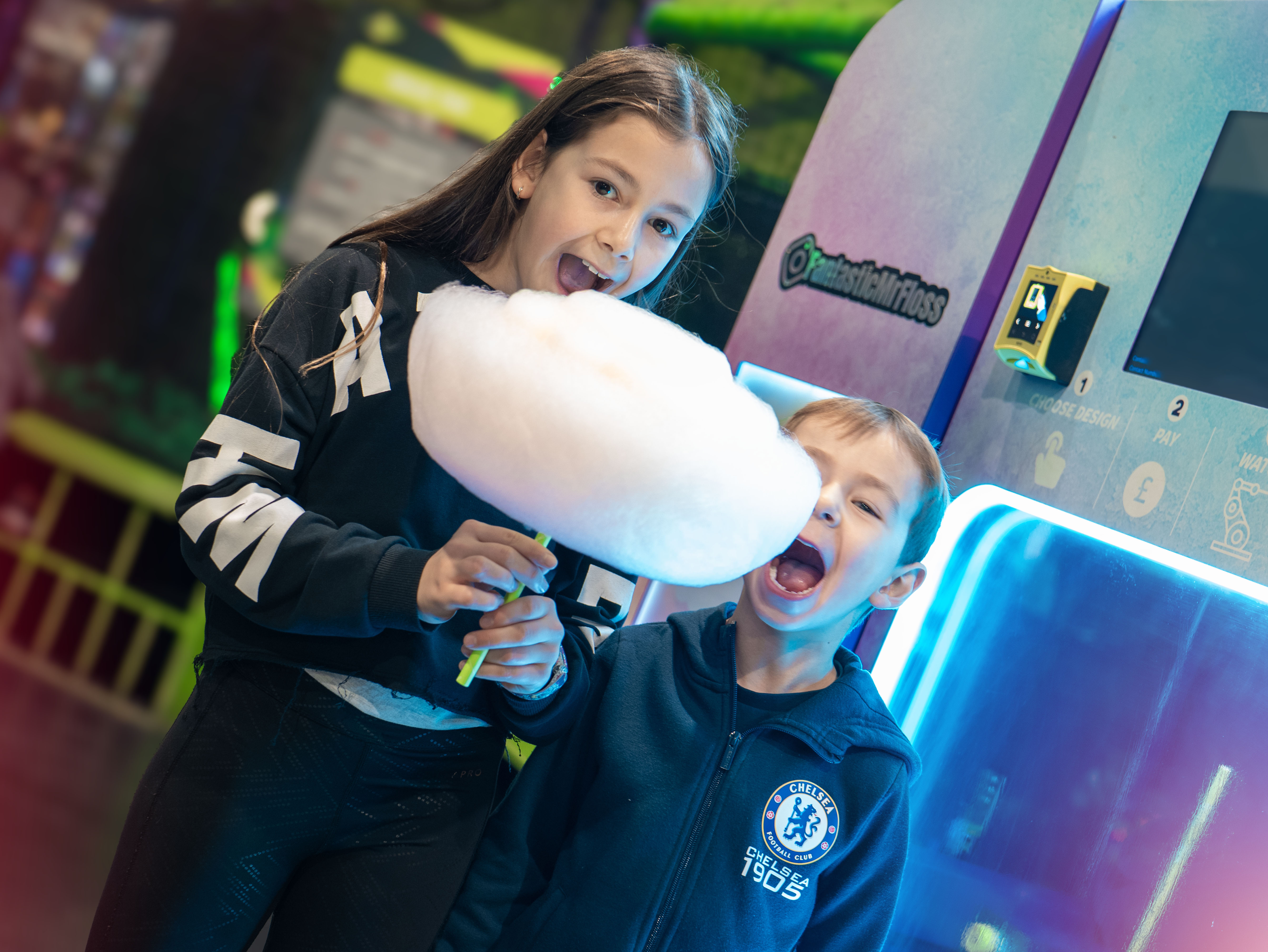 Two children excitedly enjoy a large, fluffy cotton candy inside a vibrant arcade, highlighting the fun and playful atmosphere.