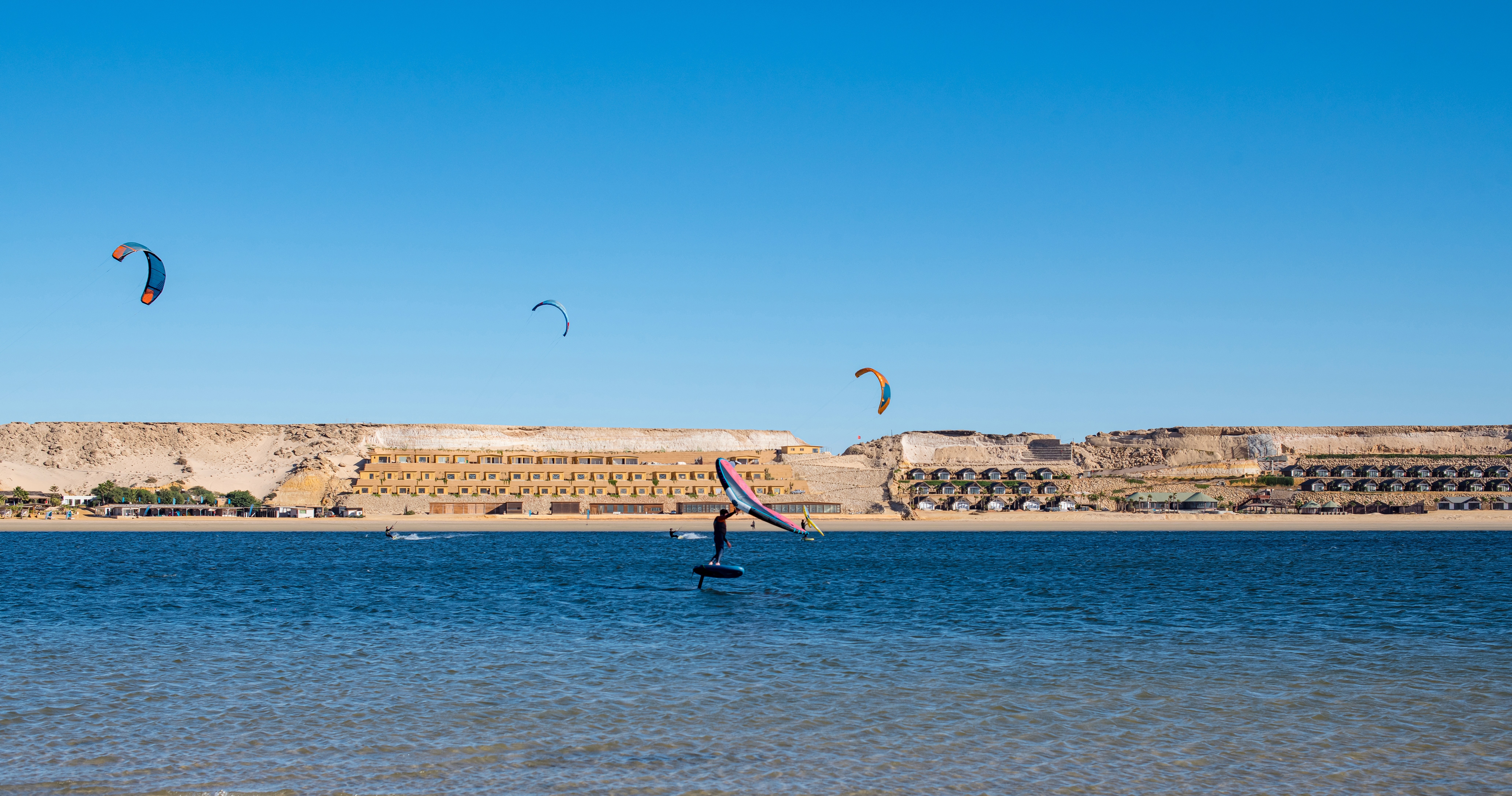A windsurfer glides across a calm blue lagoon while several colorful kitesurfers ride in the distance beneath a bright, cloudless sky.