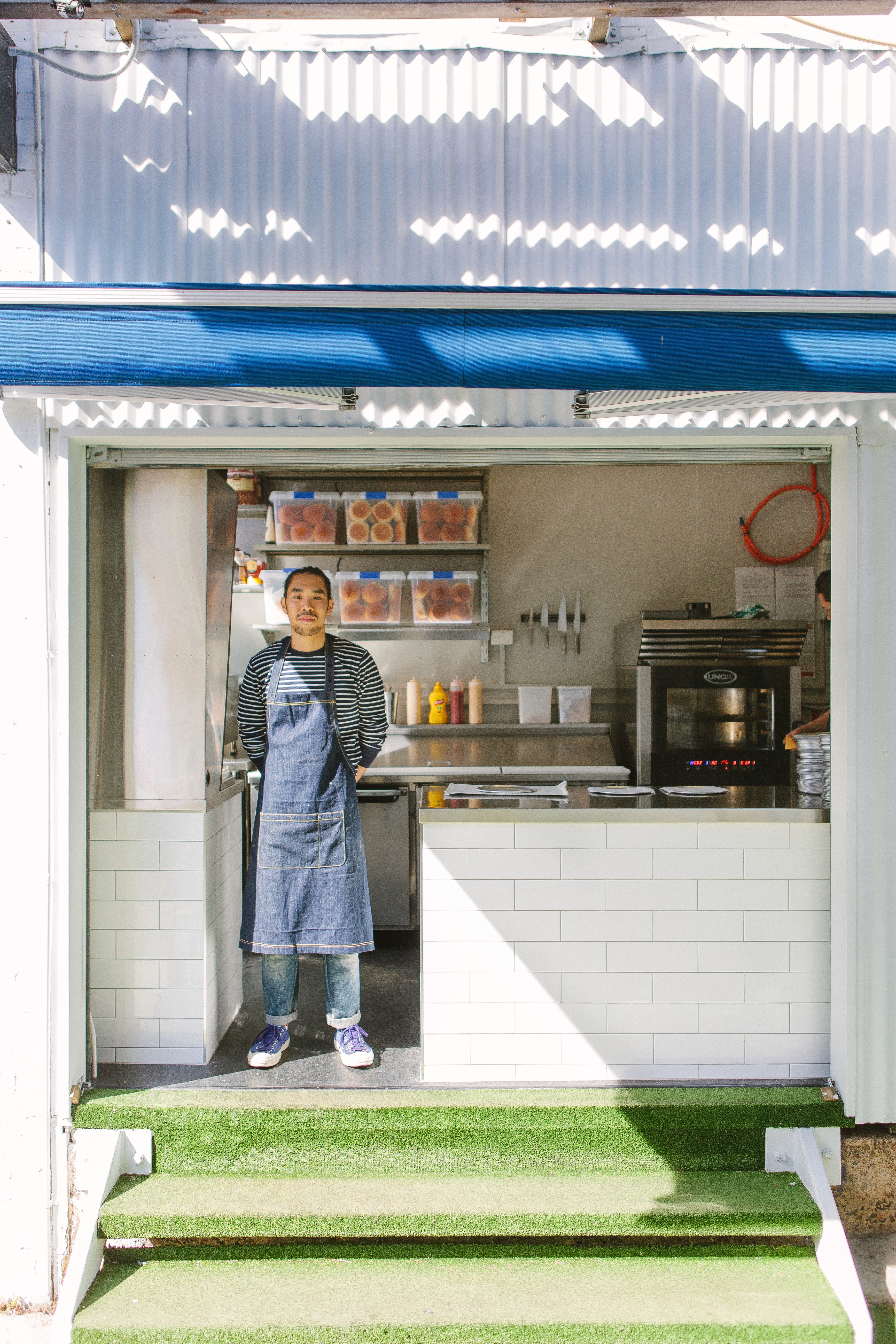 Ben’s Burgers service window with the chef standing inside the compact laneway kitchen, framed by white tiled surfaces and corrugated cladding.