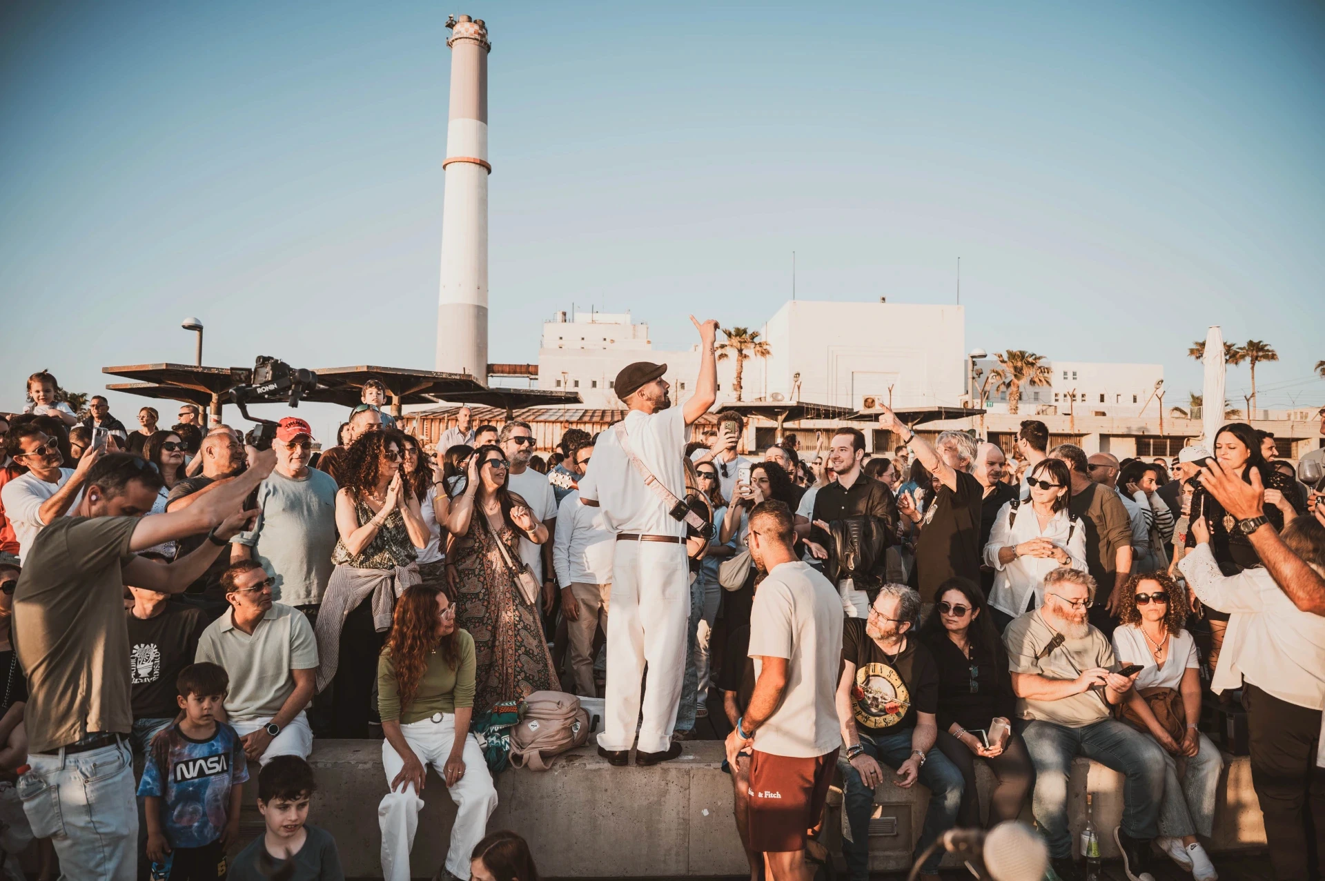 קהל בהופעה חיה בשקיעה מול תחנת הכוח ברידינג תל אביב, צילום אמנותי של הצלמת שירי עציץ Shiriz המתעד רגעי קהילה ומוזיקה תחת כיפת השמיים.