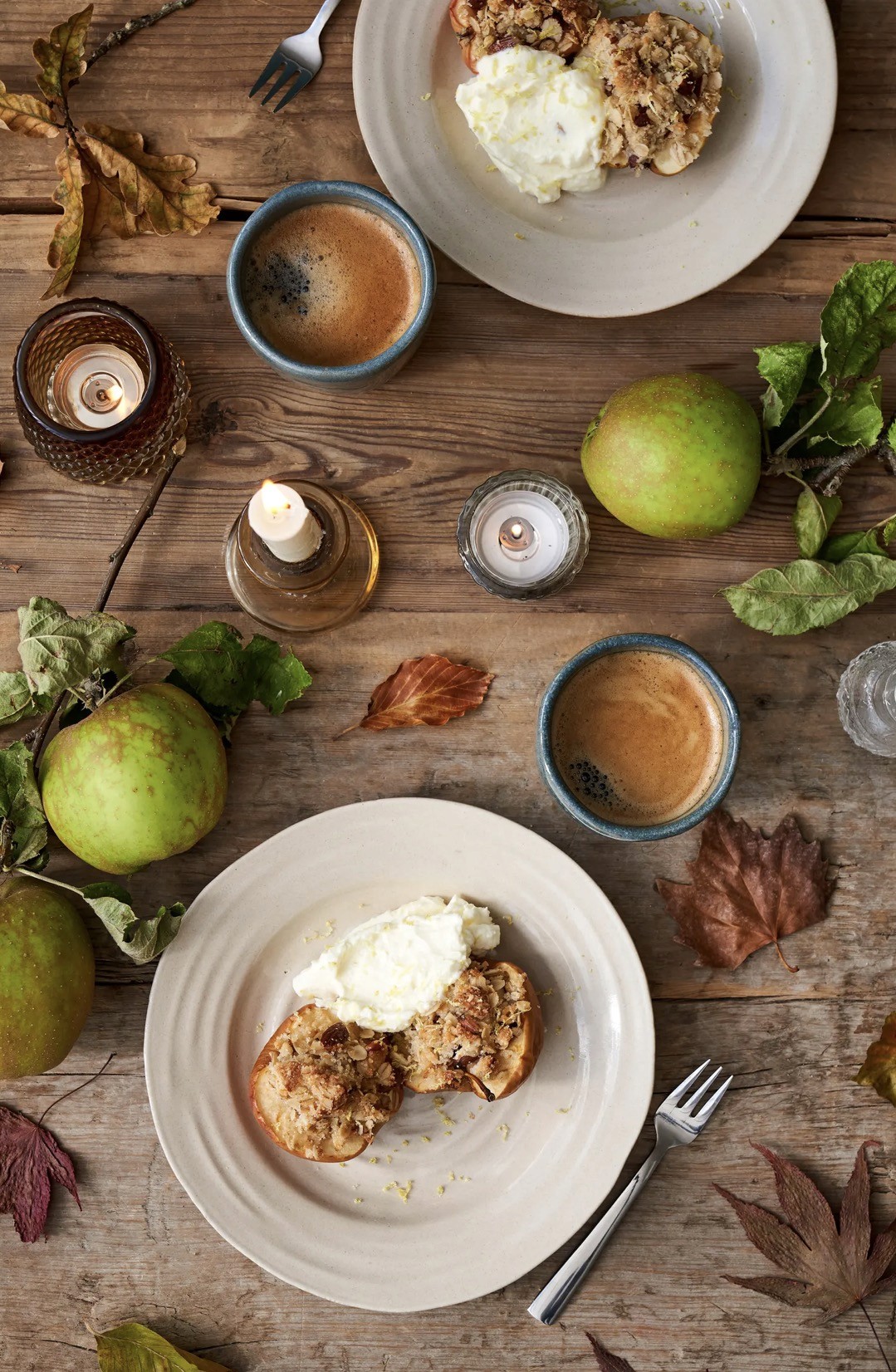 Vue du dessus de deux assiettes en grès beige avec pommes au four et granola, accompagnées de mugs à café bleus, sur table en bois avec feuilles d'automne
