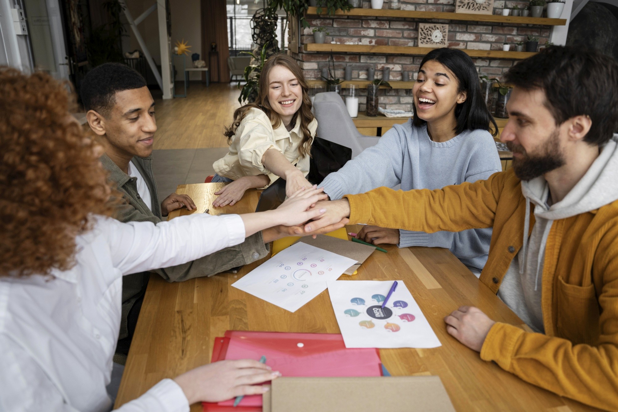 A cheerful group of colleagues stacking hands together in a gesture of teamwork around a table with documents and folders